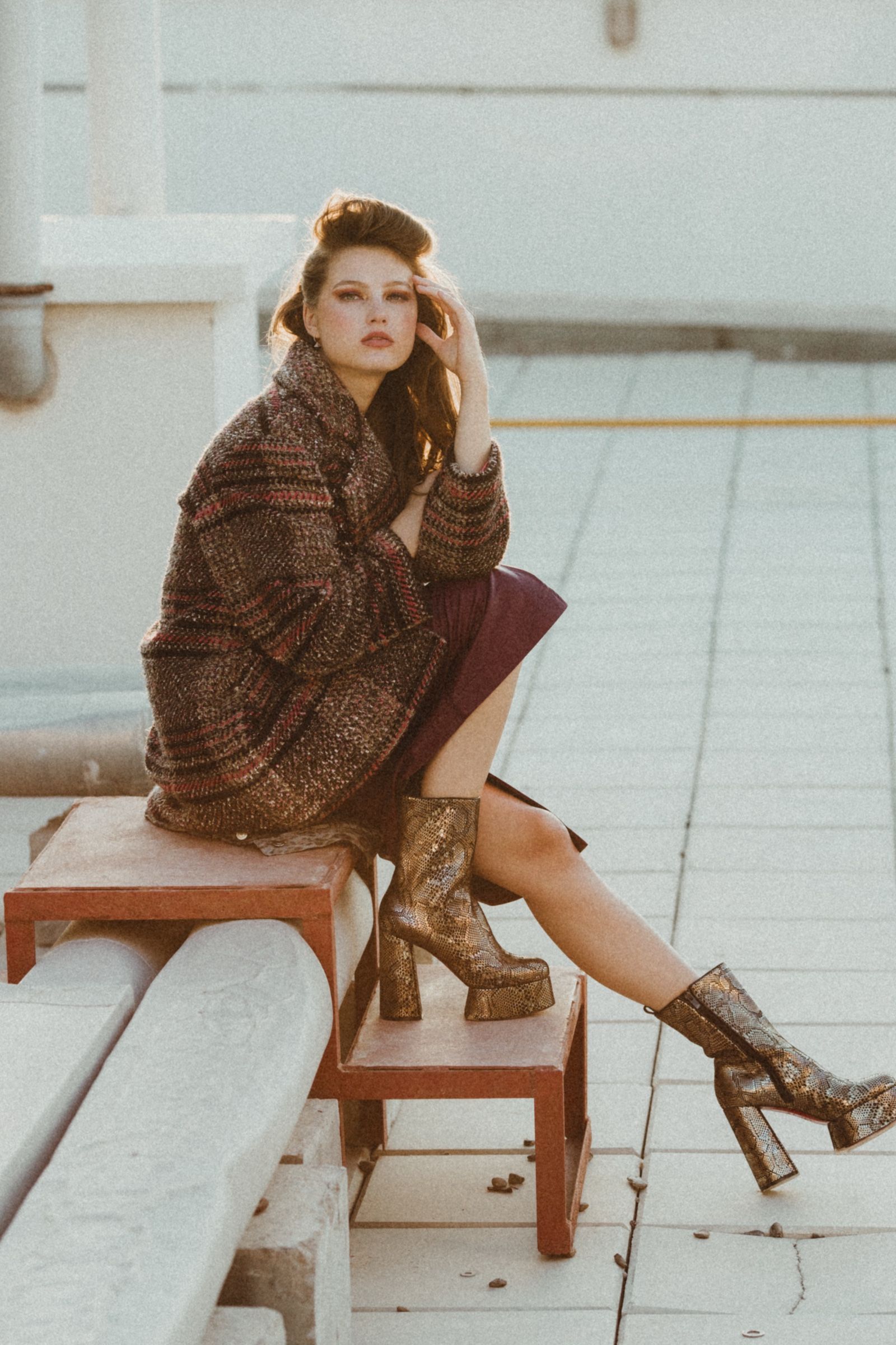 Seated on a small red bench, she wears a heavy striped knit coat over a plum skirt with tall snakeskin platform boots, grainy warm light.