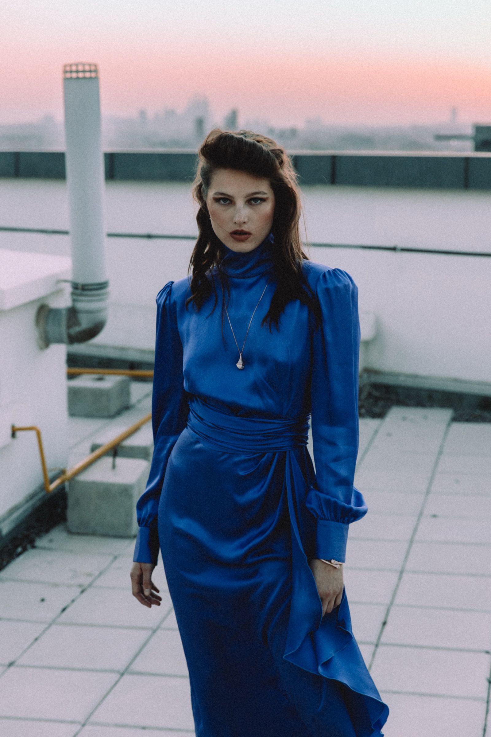 The model stands on a white rooftop deck in a cobalt satin gown with high neck, gathered sleeves and wrapped waist, skyline pink behind her.