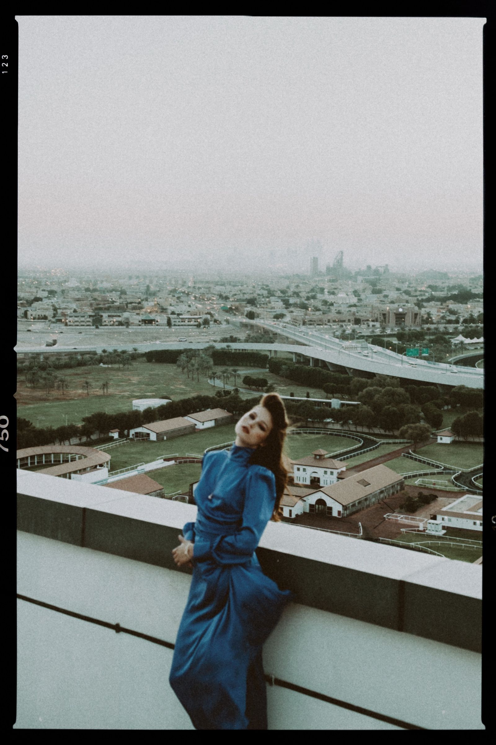 Grainy frame with the film rebate showing, the model reclining against a rooftop parapet in the blue satin gown above a hazy grey city at dusk.