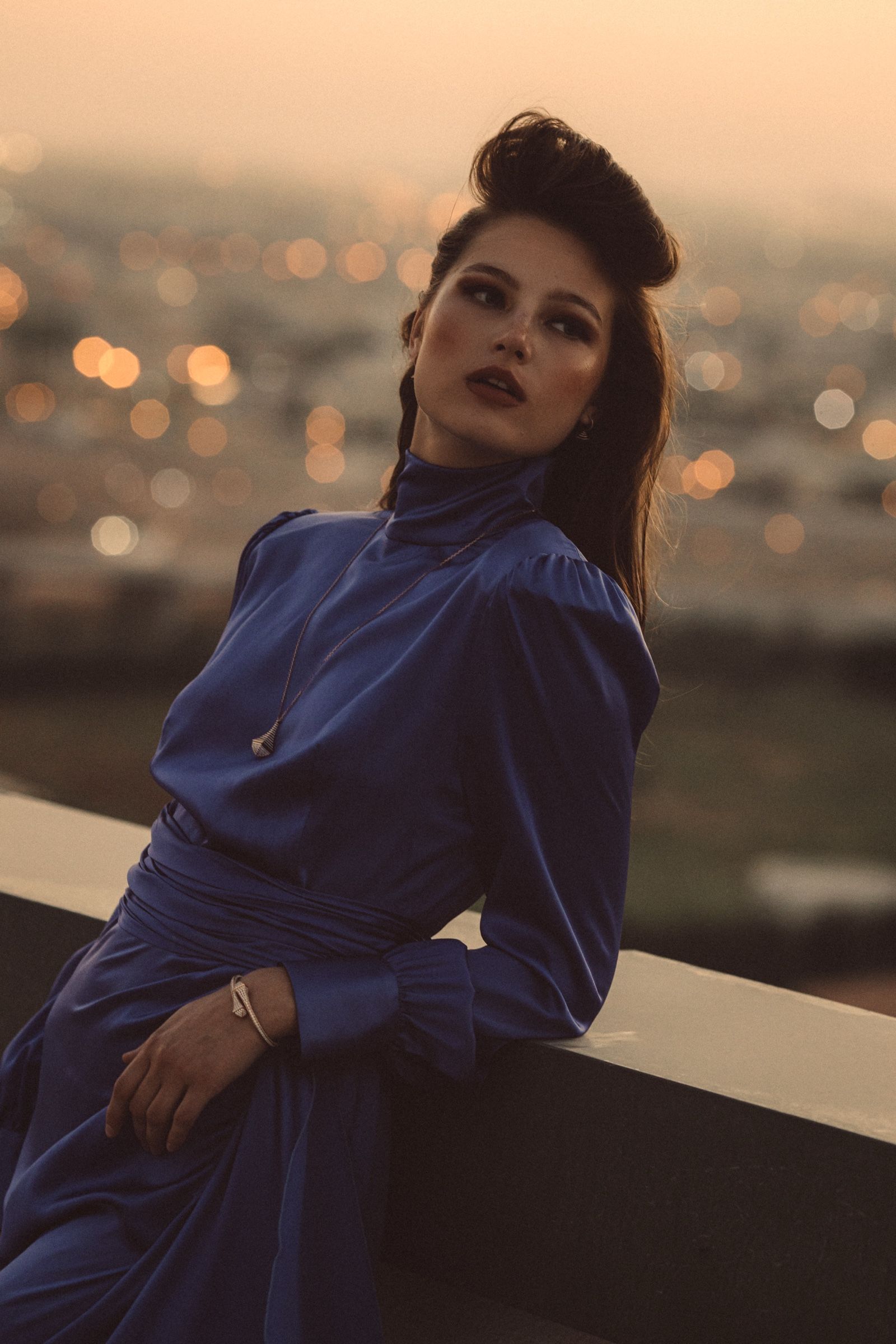 Grainy close frame of the model leaning on a rooftop ledge in the blue satin gown, a fine pendant chain at her neck, city lights blurred gold behind.