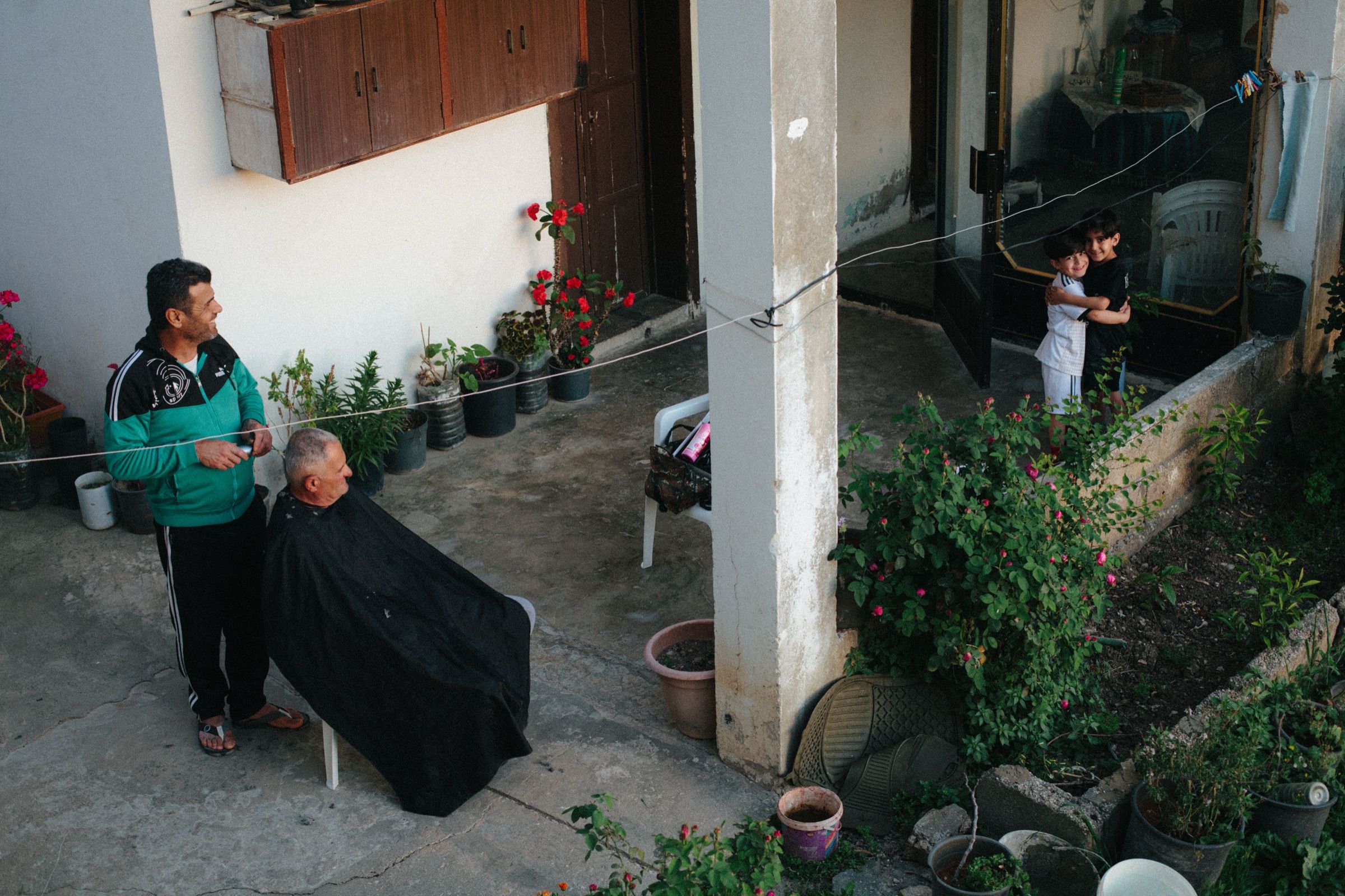 A man receives a haircut from a local barber in the courtyard garden of a modest home at dusk. Rows of potted plants line the perimeter. In the background, two children, distant relatives of the barber, embrace near the entrance, looking toward the camera. The town of Joun, Lebanon, 2025.