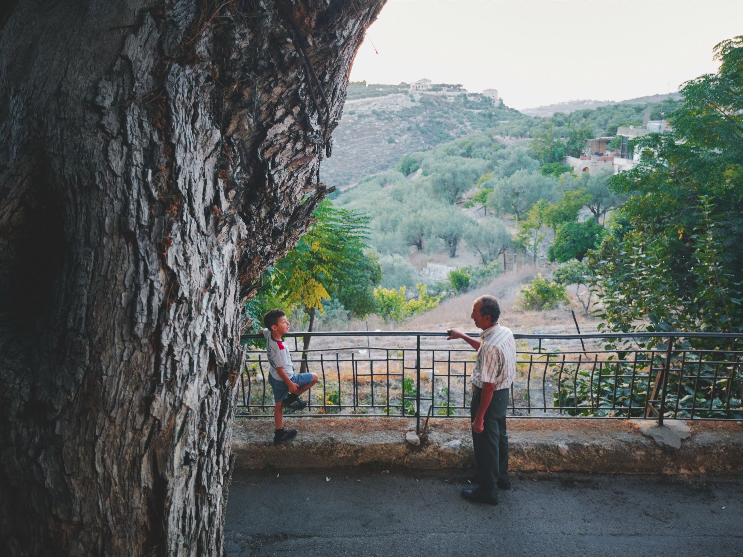 A grandfather and his grandson rest their hands on a steel railing at dusk, talking to each other in one of Joun’s neighborhoods, with the village’s mountains stretching out in front of them. The town of Joun, Lebanon, 2025.