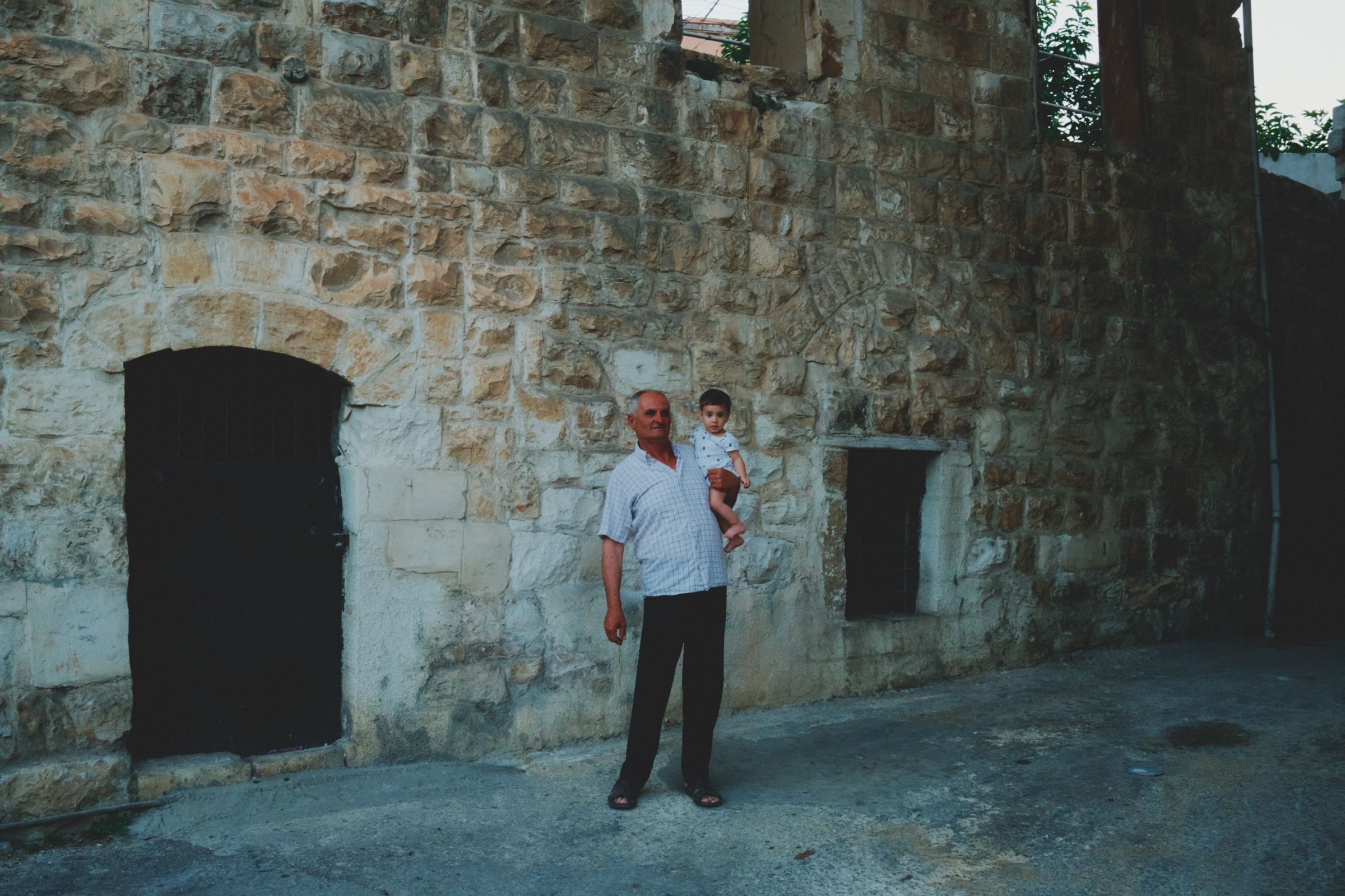 A grandfather holds his grandson near their home in one of the oldest village alleys, 2024.