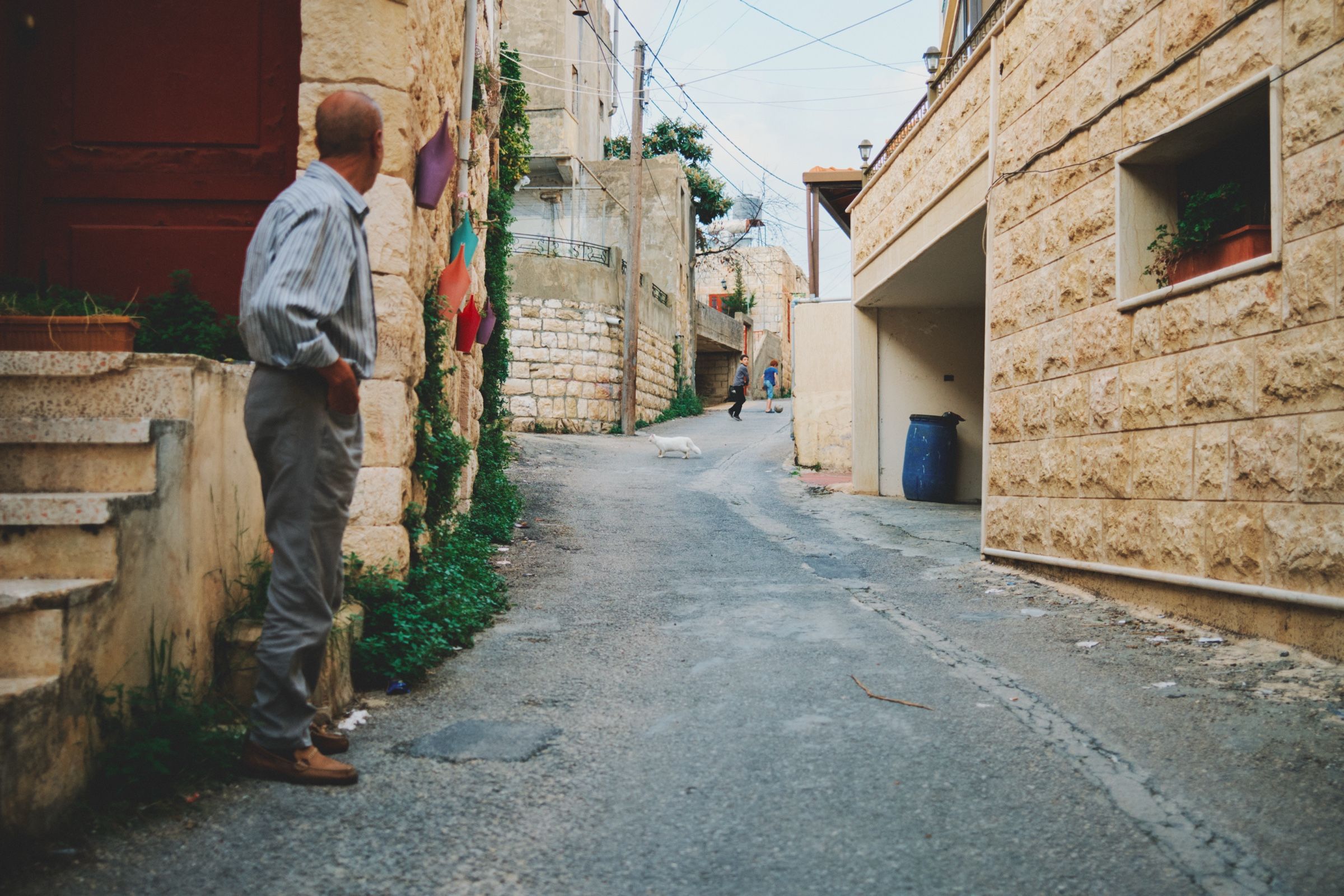 An old man is watching two kids run through a narrow street in the village of Joun, as a white cat passes by. The town of Joun, Lebanon, 2023.