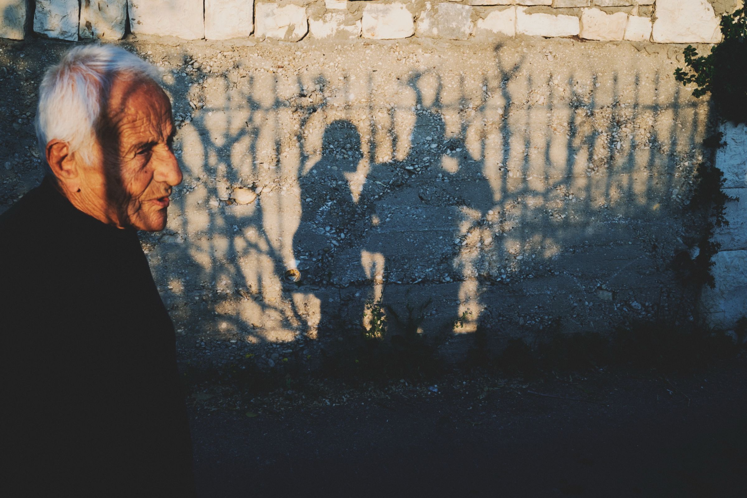 An elderly man walks along a stone wall as the photographer’s shadow falls beside him in Joun. The town of Joun, Lebanon, 2025.