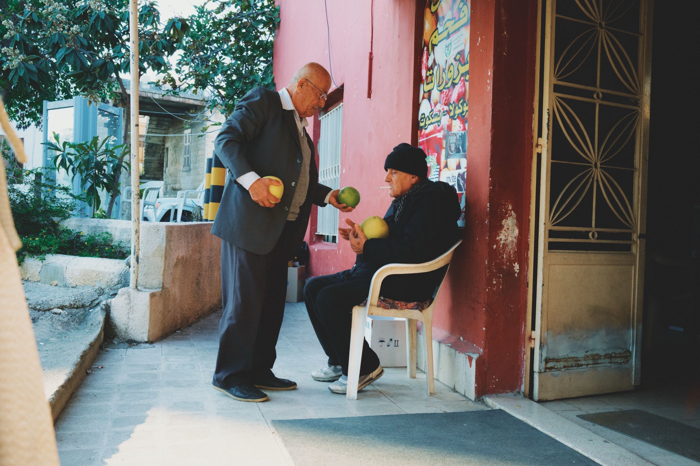 Two elderly men, one handing grapefruits to the other, 2023.