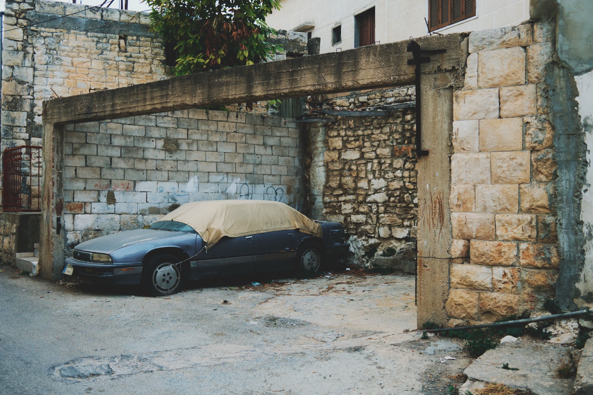 A covered, rusted car sits in a weathered garage in Joun, surrounded by old walls and overgrown plants. The town of Joun, Lebanon, 2025.
