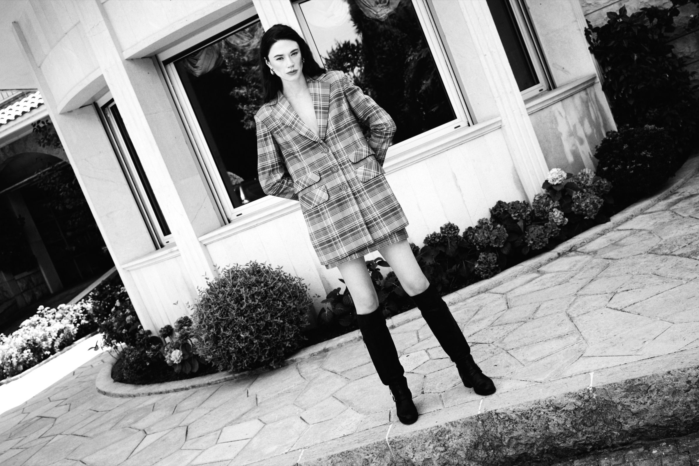 In black and white, a model in an oversized checked coat and tall boots stands on a flagstone terrace, hands behind her back.