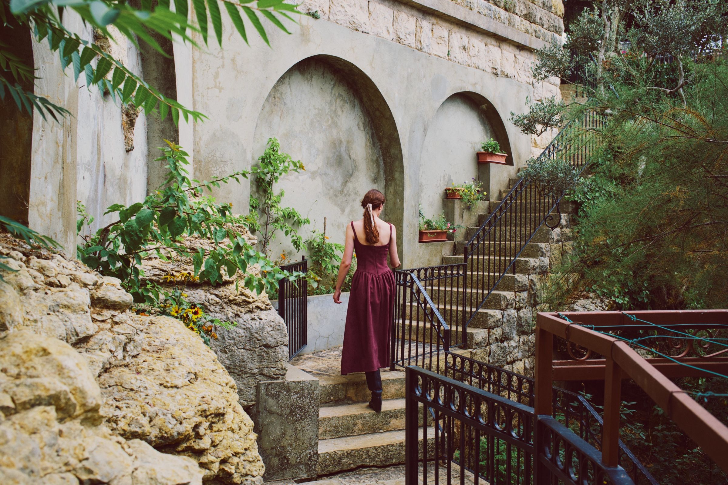 Seen from behind, the model climbs stone steps in a burgundy dress with narrow straps and a gathered skirt, past arched plaster walls and potted plants.