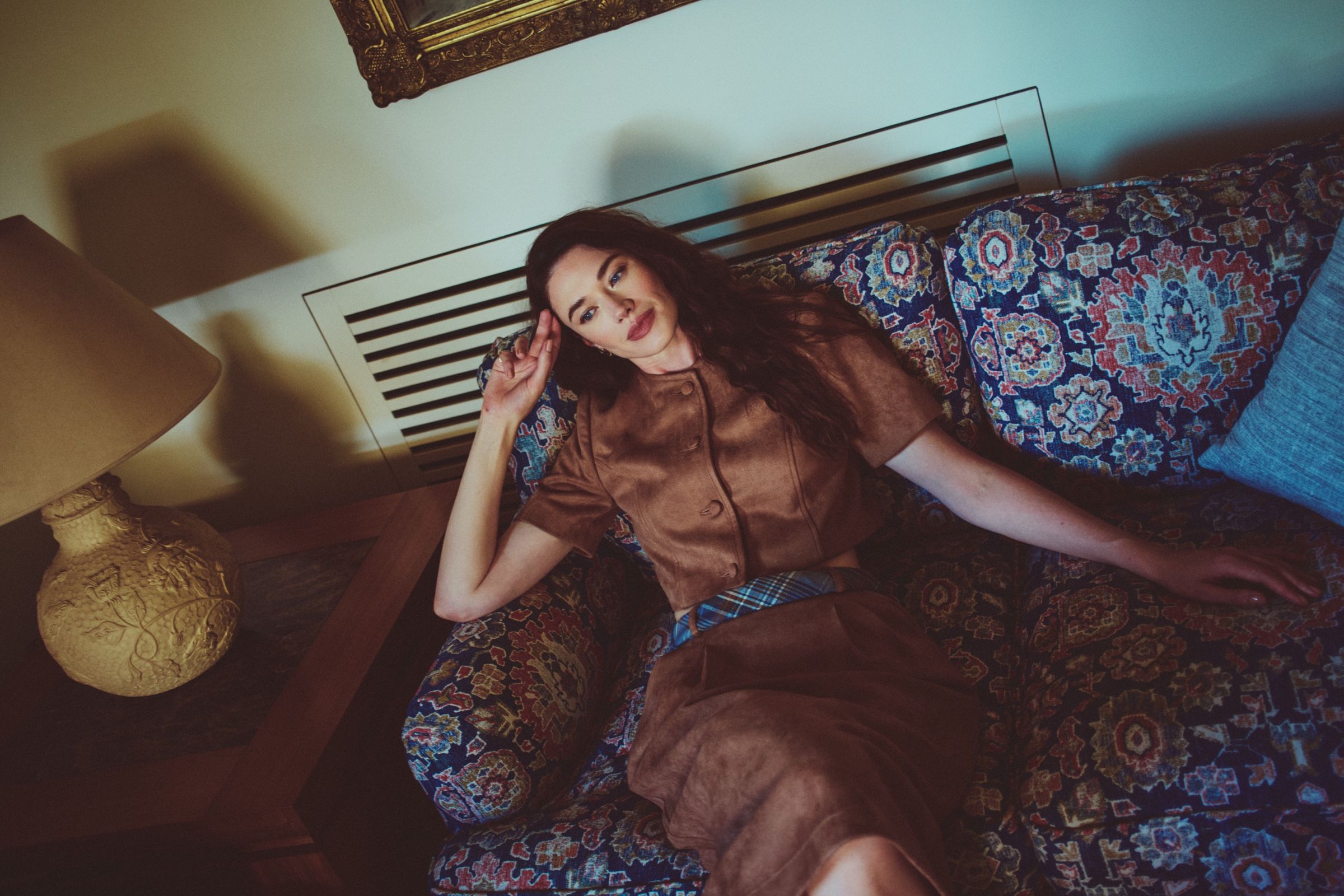 The model lies back across a patterned tapestry sofa in a short-sleeved brown suede jacket and matching skirt, a table lamp lit beside her.