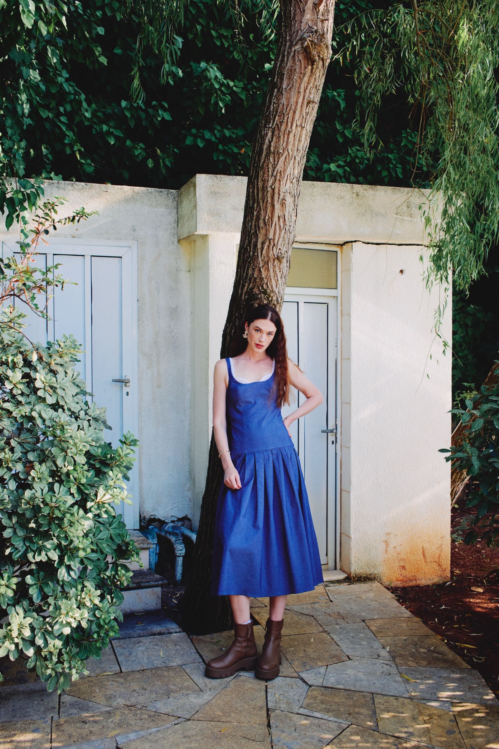 Hand on hip beside a tree trunk on flagstone paving, the model wears a denim drop-waist dress with lace trim and brown platform boots.