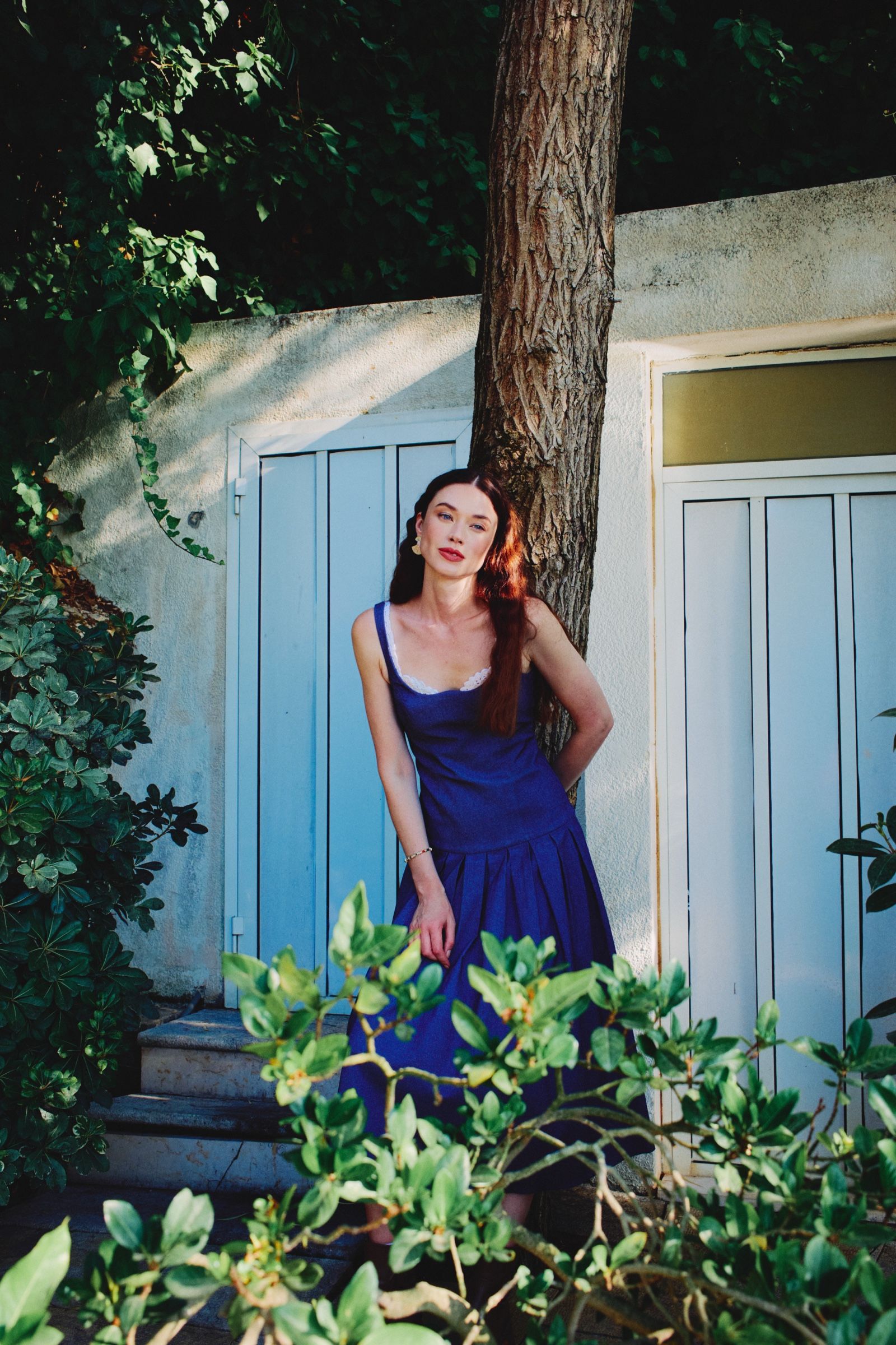 The model stands beside a tree trunk in a sleeveless denim dress, pale blue panelled doors and dappled foliage behind her.