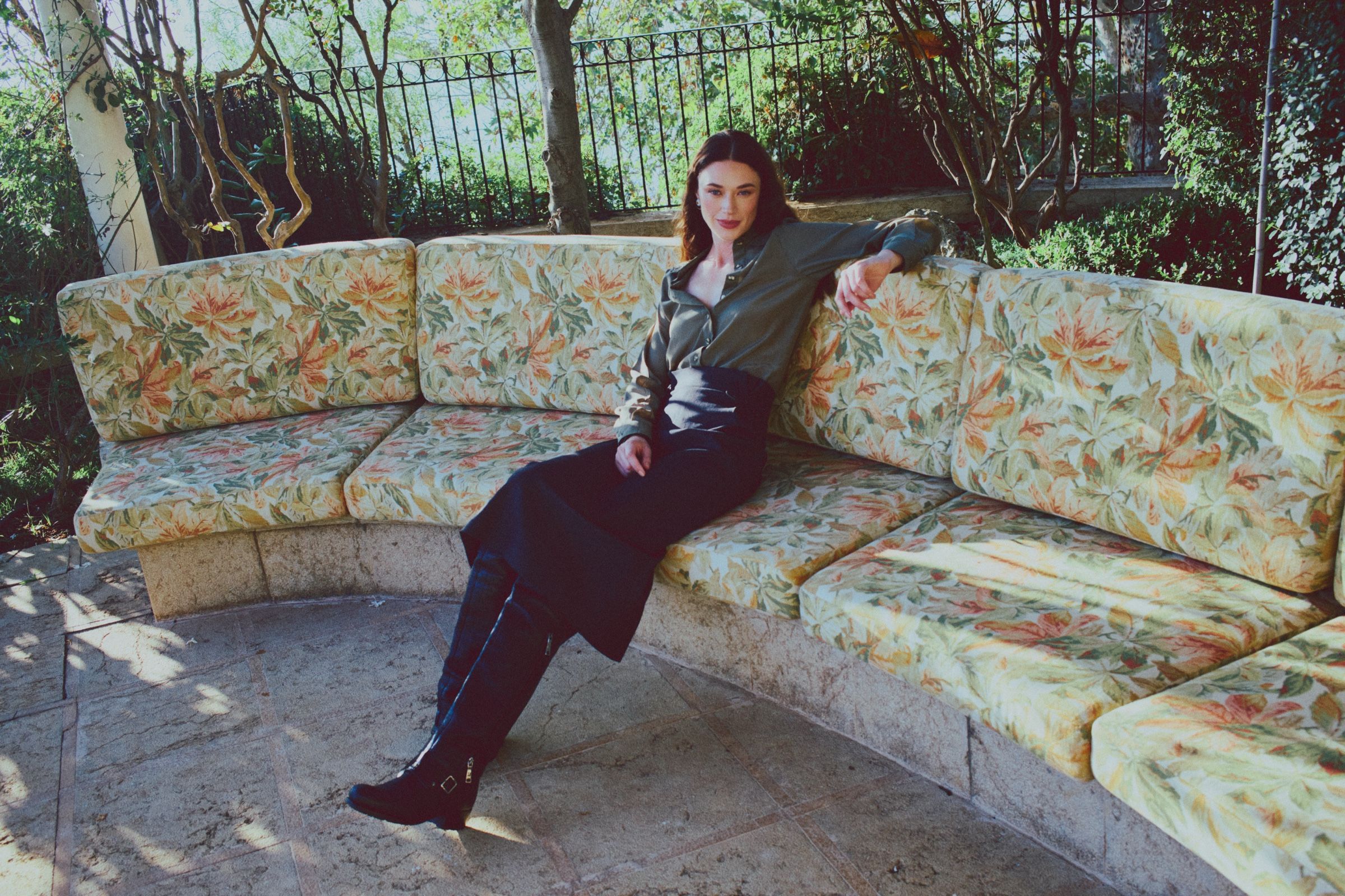 The model sits back on a curved banquette of leaf-print cushions, one arm along the backrest, in an olive satin shirt and tall black boots.