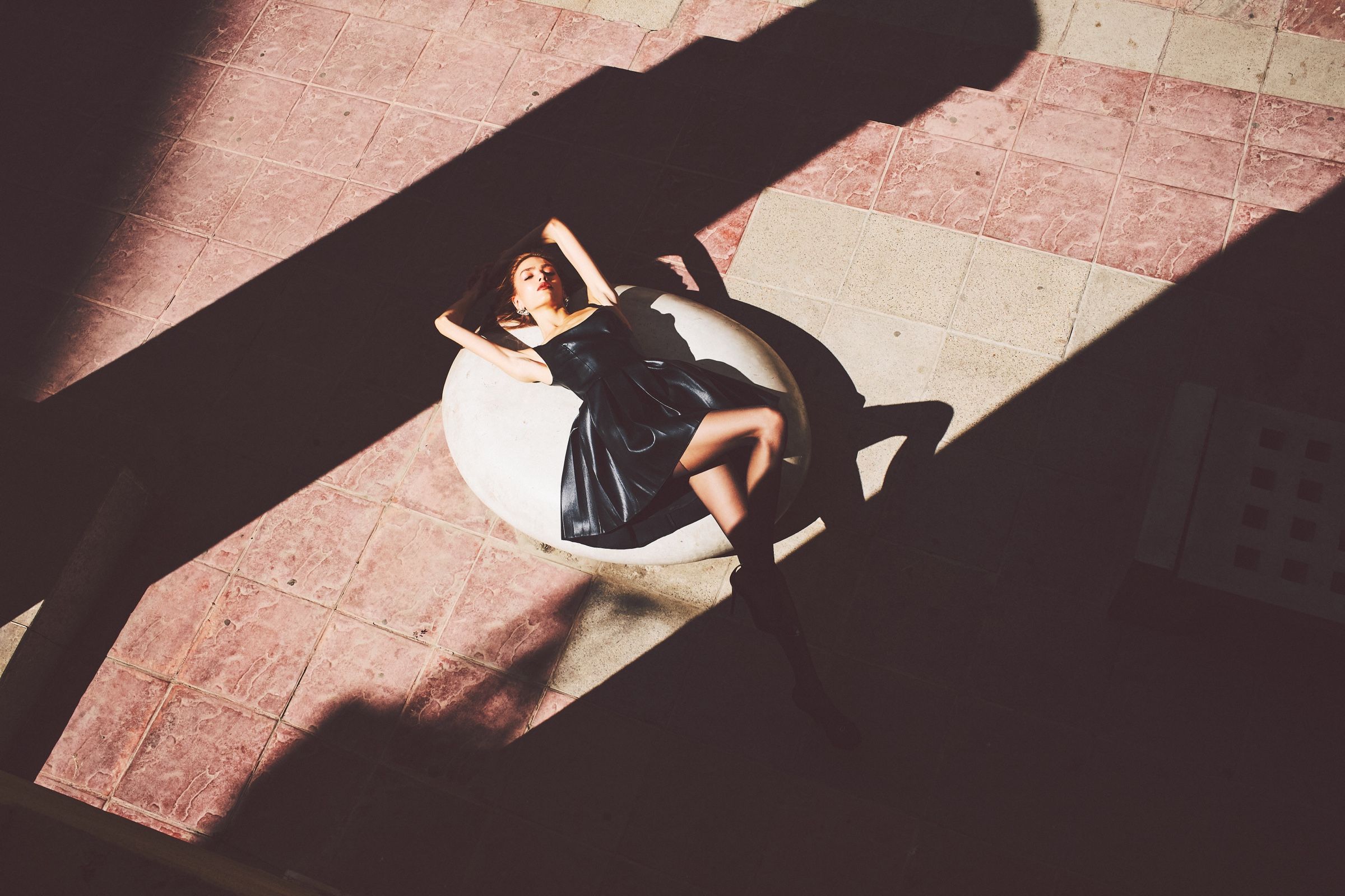 Seen from above, a model in a black leather minidress and knee boots reclines on a white stone sphere across a hard diagonal shadow.