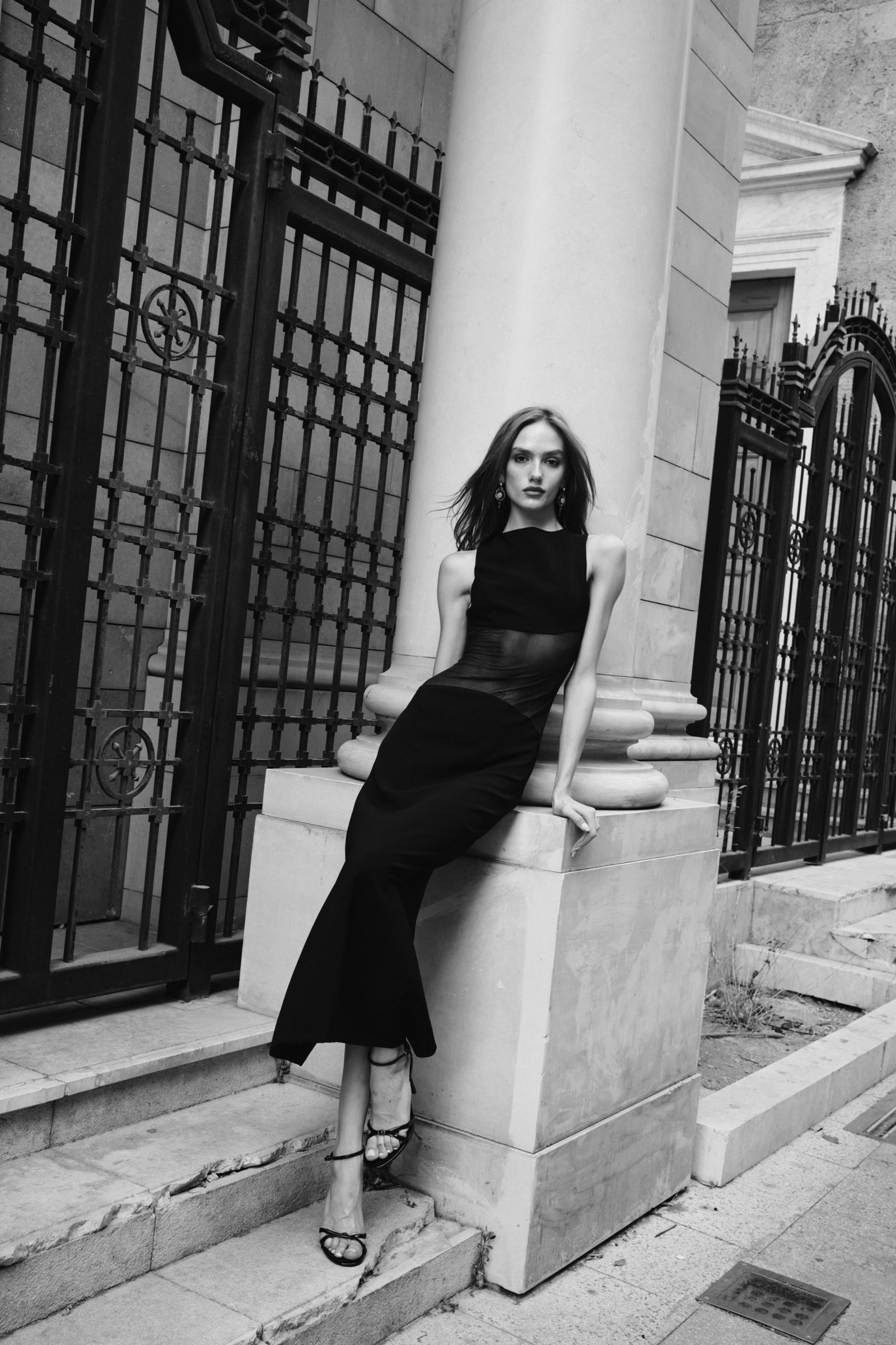 In black and white, a model in a sleeveless black gown with a sheer waist panel sits on a column plinth beside iron railings.
