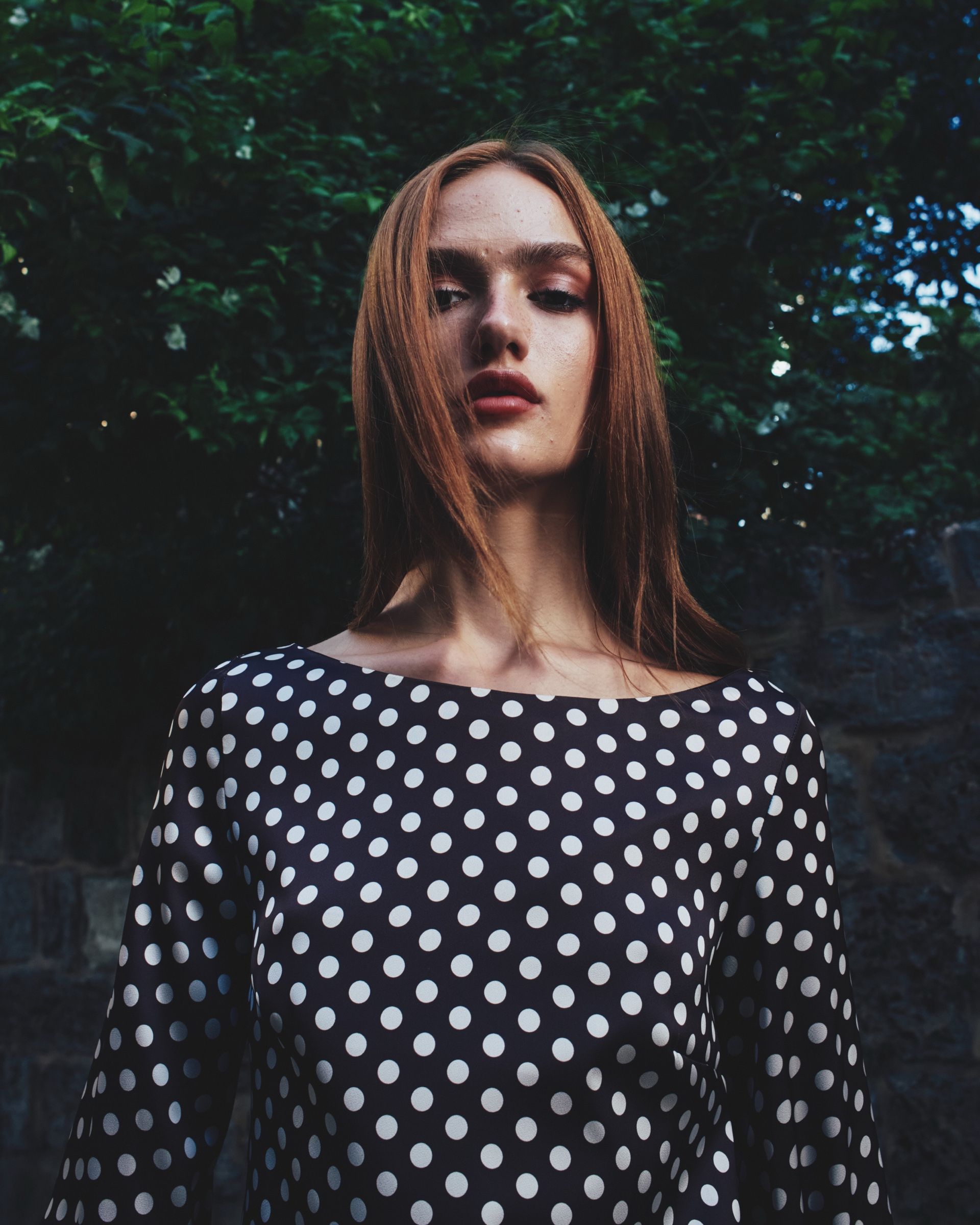 Shot from below, a model with long auburn hair in a navy polka-dot top stands against dark foliage and a stone wall.