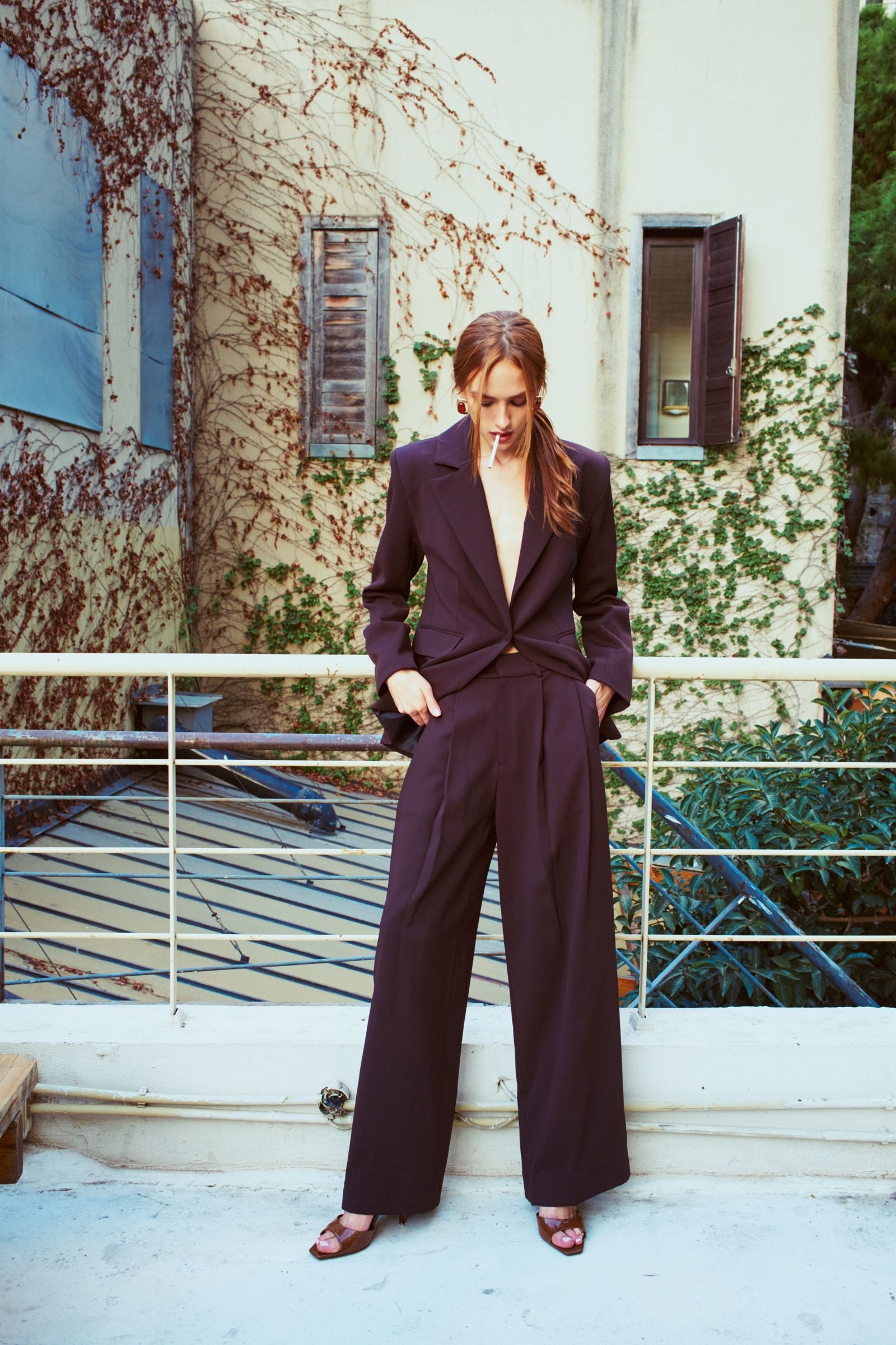 A model in a deep aubergine suit with wide trousers looks down, cigarette in mouth, against an ivy-covered wall.