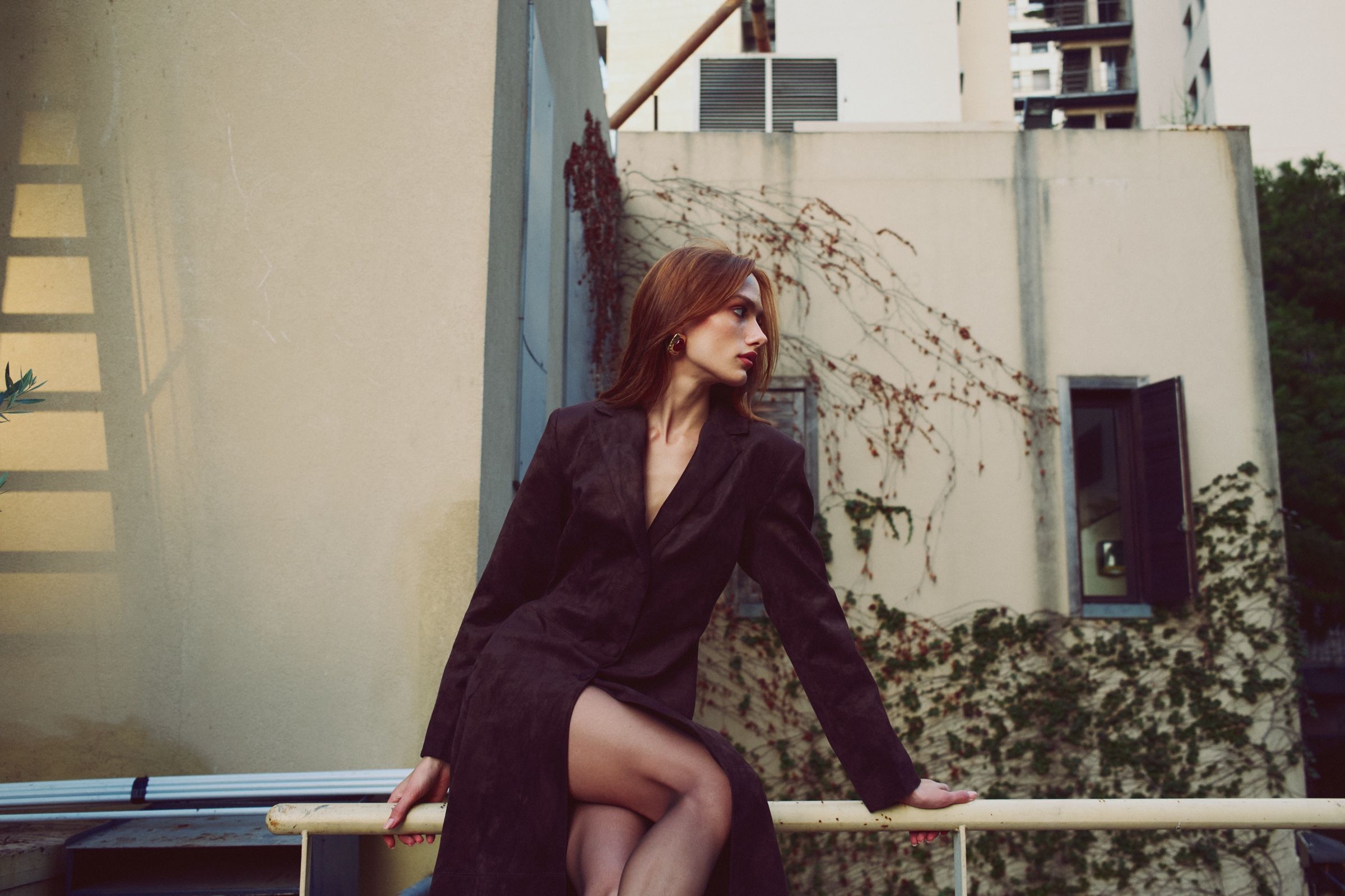Seated on a railing in a deep-V suede coat, the model turns her face into profile, pale rooftop walls behind her.