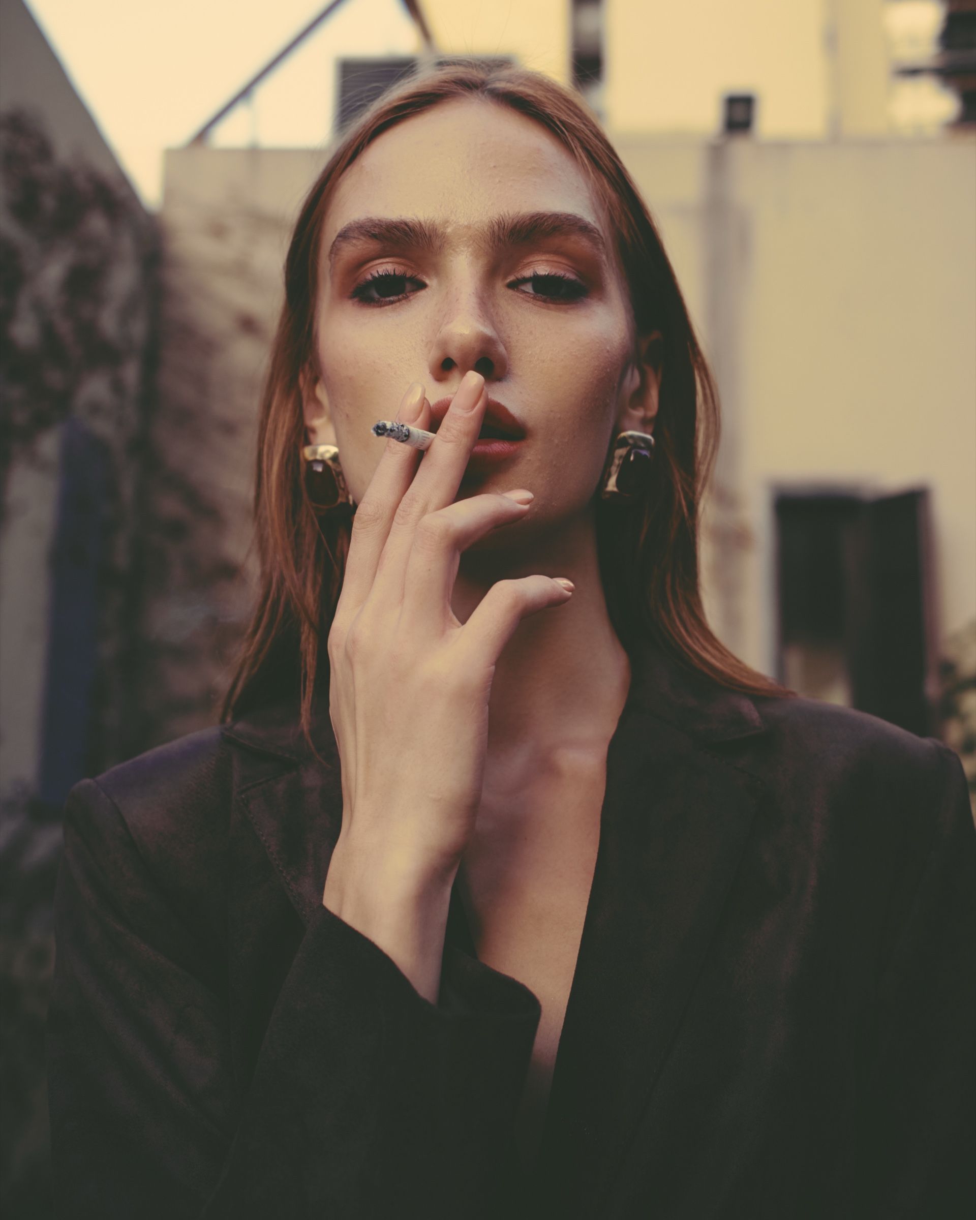 Close portrait of a model in a dark blazer and gold-set earrings, holding a cigarette to her lips in warm evening light.