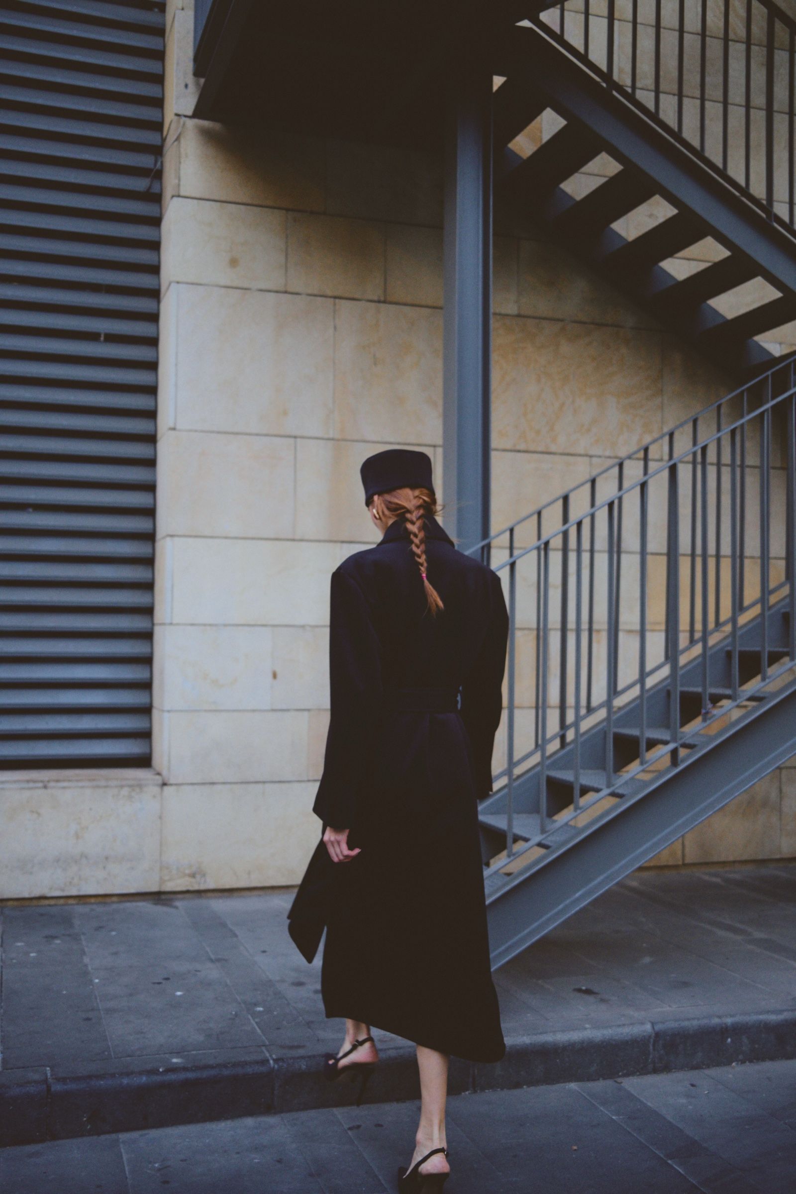 Seen from behind, a model in a belted dark coat and flat-topped hat walks past a limestone wall, a braid down her back.