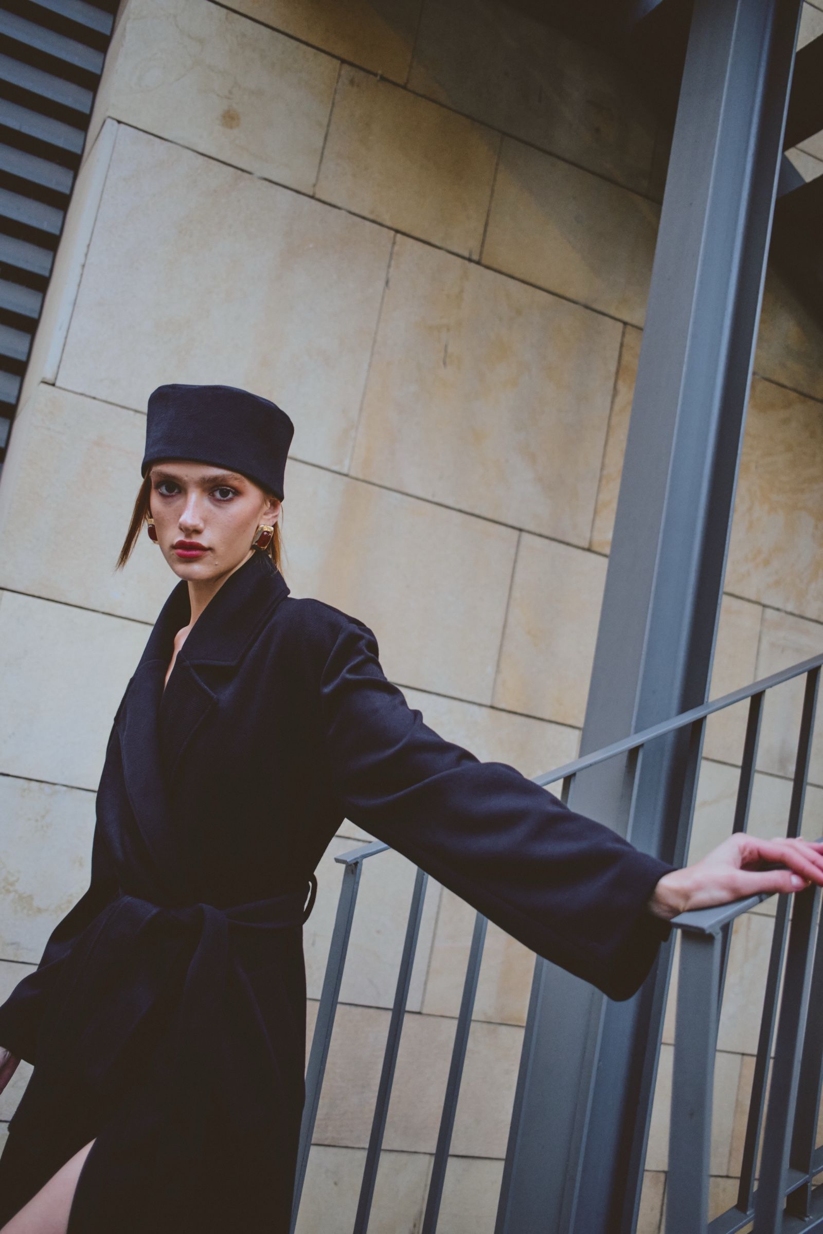 A model in a belted navy wool coat and matching flat-topped hat rests one arm along a steel railing against a limestone wall.