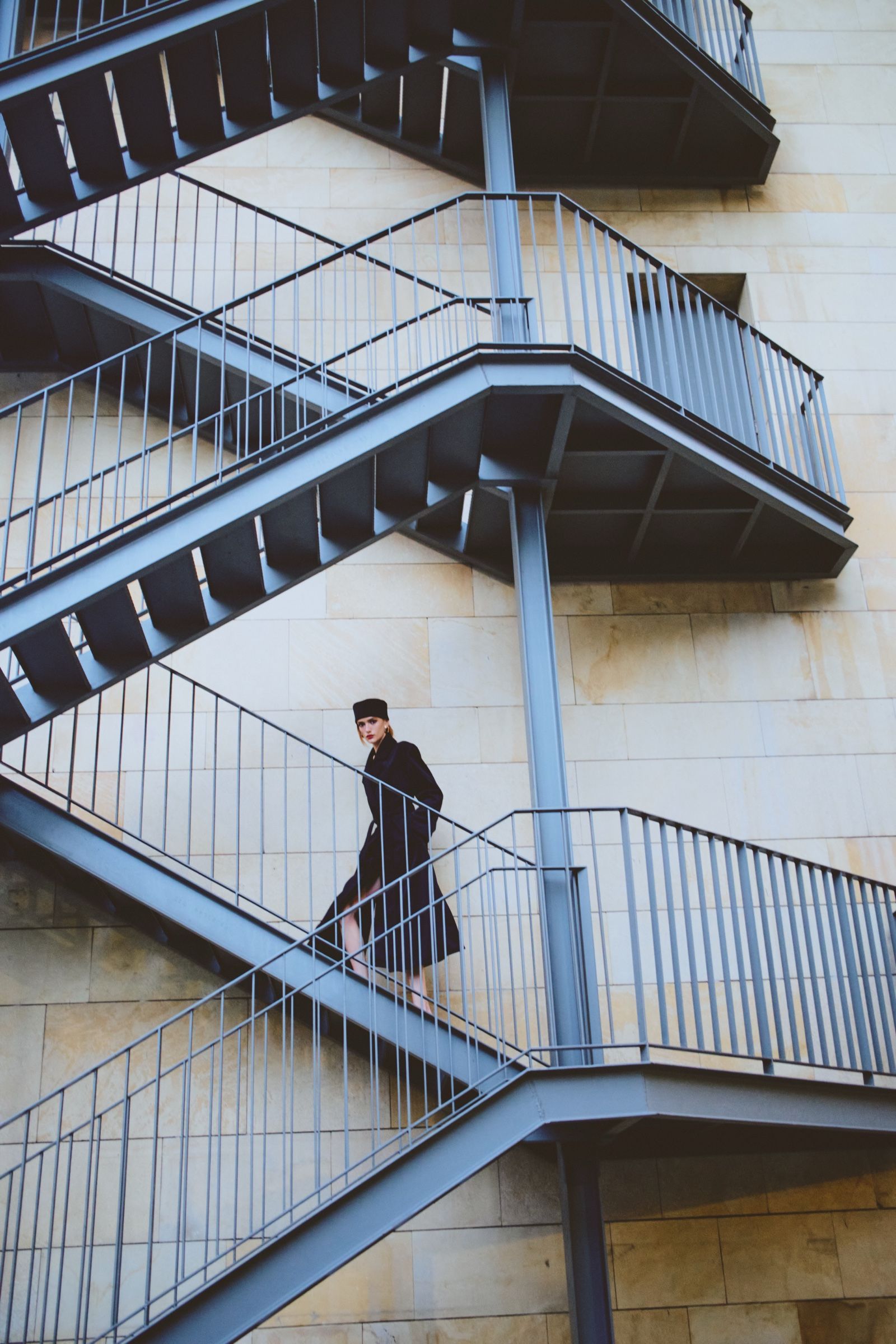 Small in the frame, a model in a long dark coat and flat-topped hat pauses on a landing of a grey steel fire escape.