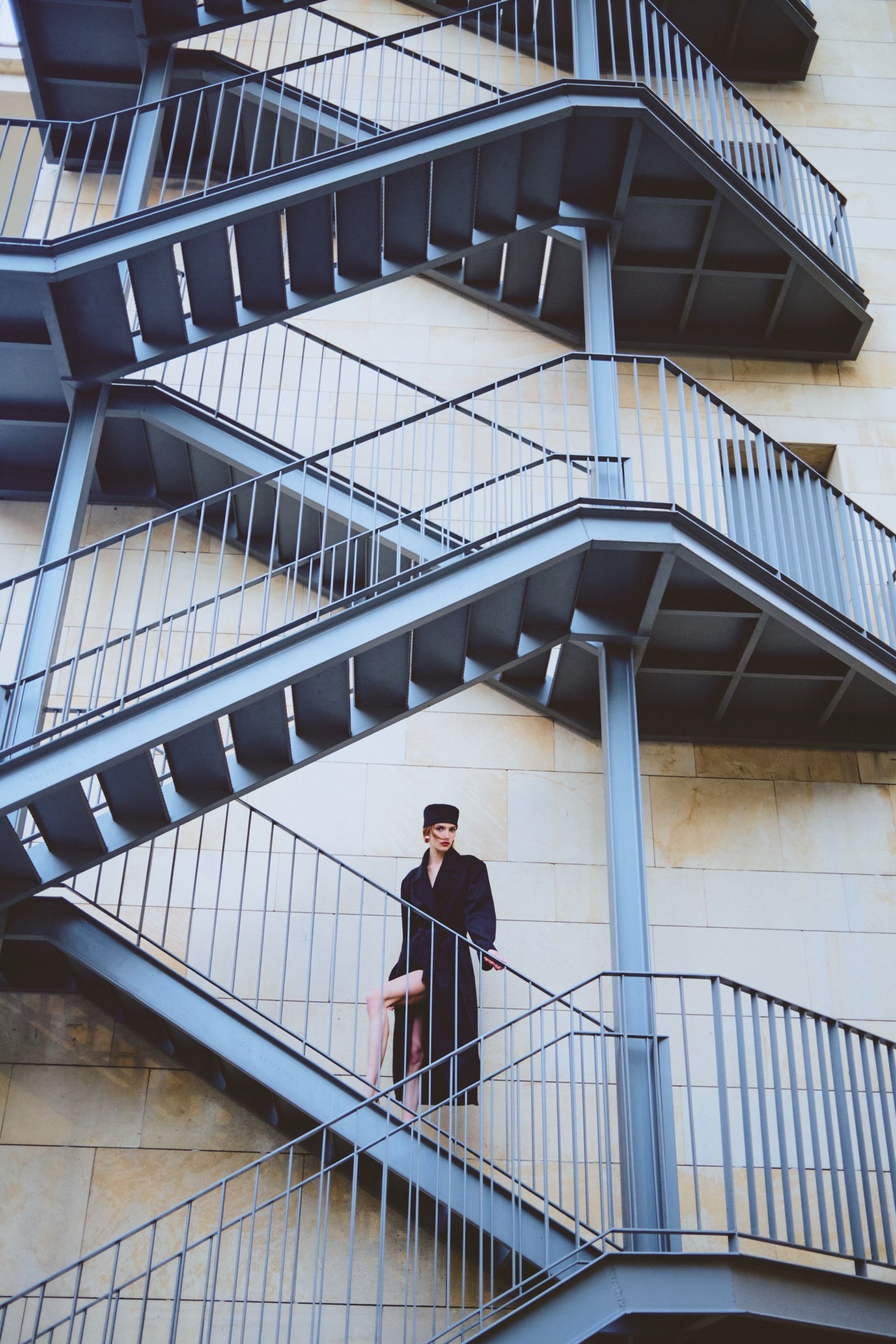 Mid-flight on a zigzagging steel fire escape, a model in a long dark coat and pillbox hat steps forward, one leg bare.