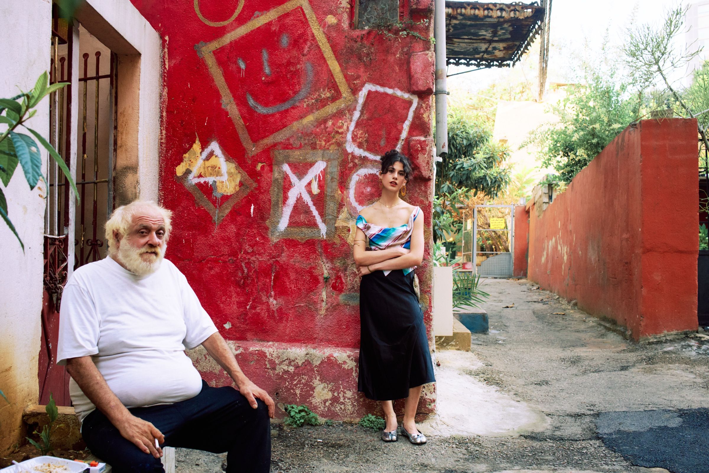 A model in a striped satin top and long black skirt stands arms folded against a red graffitied wall, a bearded man seated nearby.