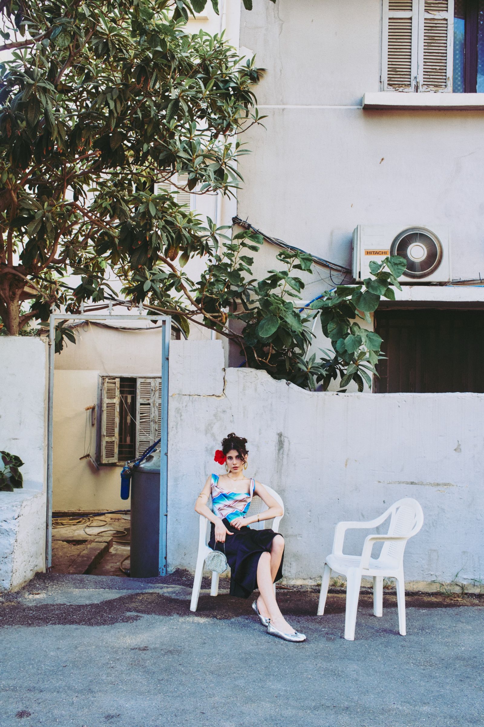The model sits with legs crossed on a white plastic chair, small in a wide frame of whitewashed walls, shutters and overhanging branches.