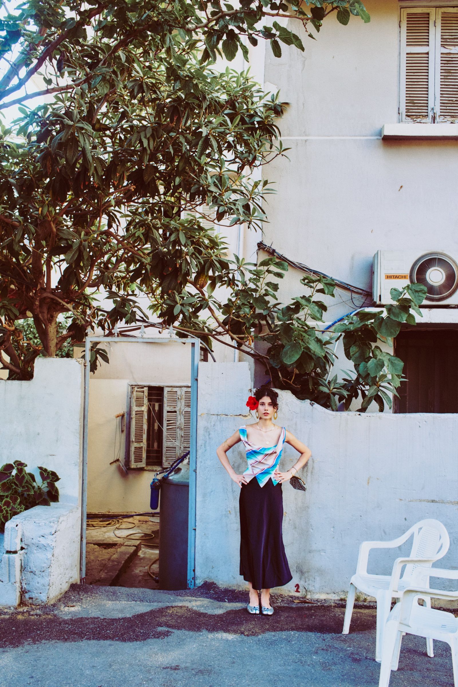 Hands on hips, the model stands before an open metal gate in a long black satin skirt, stacked plastic chairs at right.