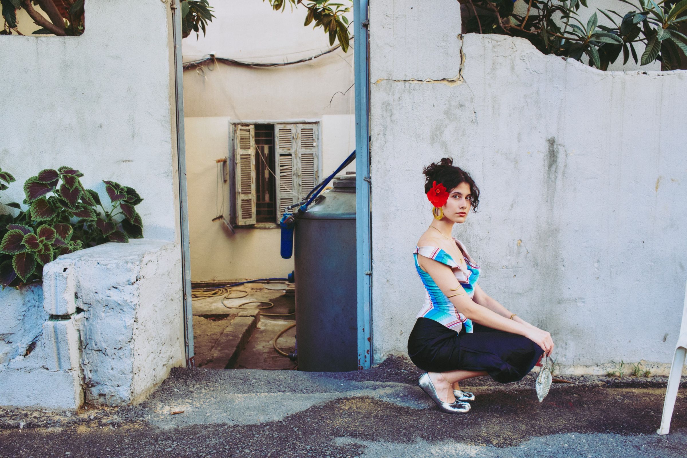 The model crouches on her heels beside a pale concrete wall, a small silver beaded purse hanging from one hand.