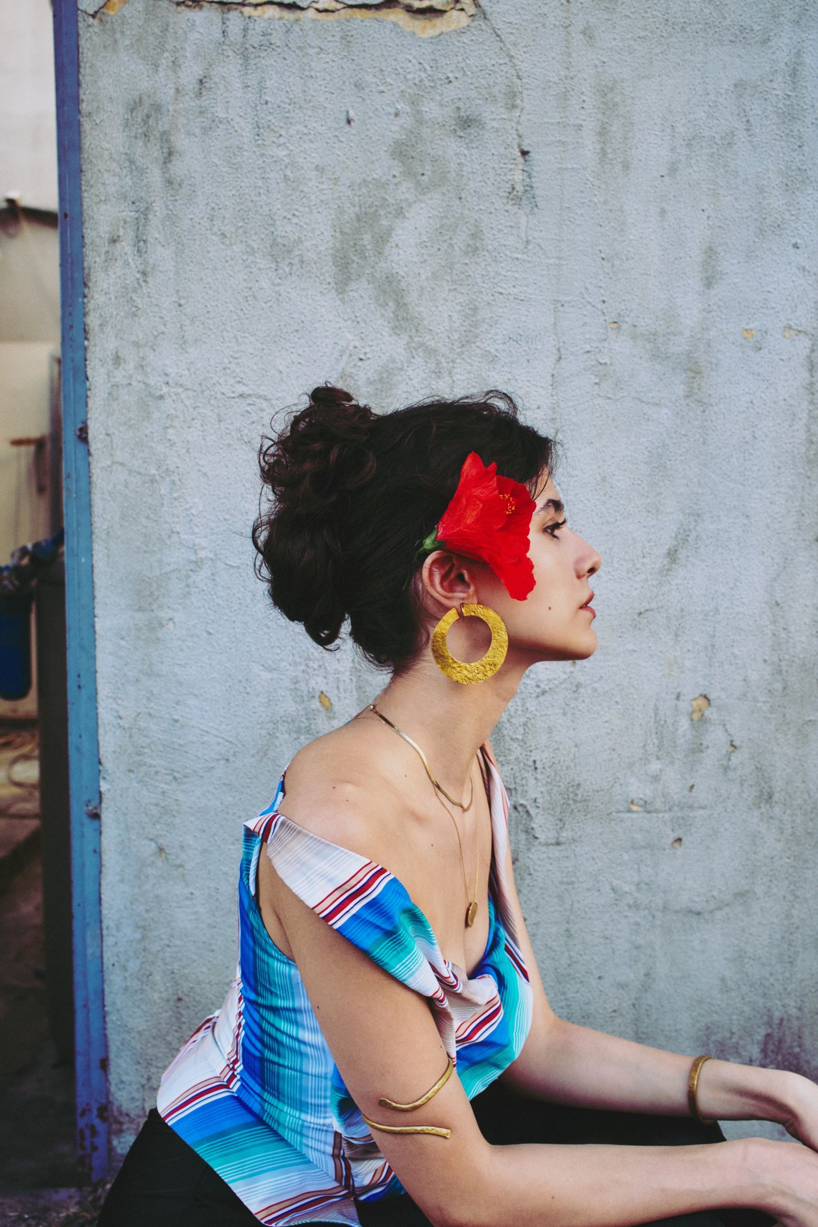 Close profile of the model, a red hibiscus at her ear, hammered gold hoop swinging, the striped satin top slipping off one shoulder.