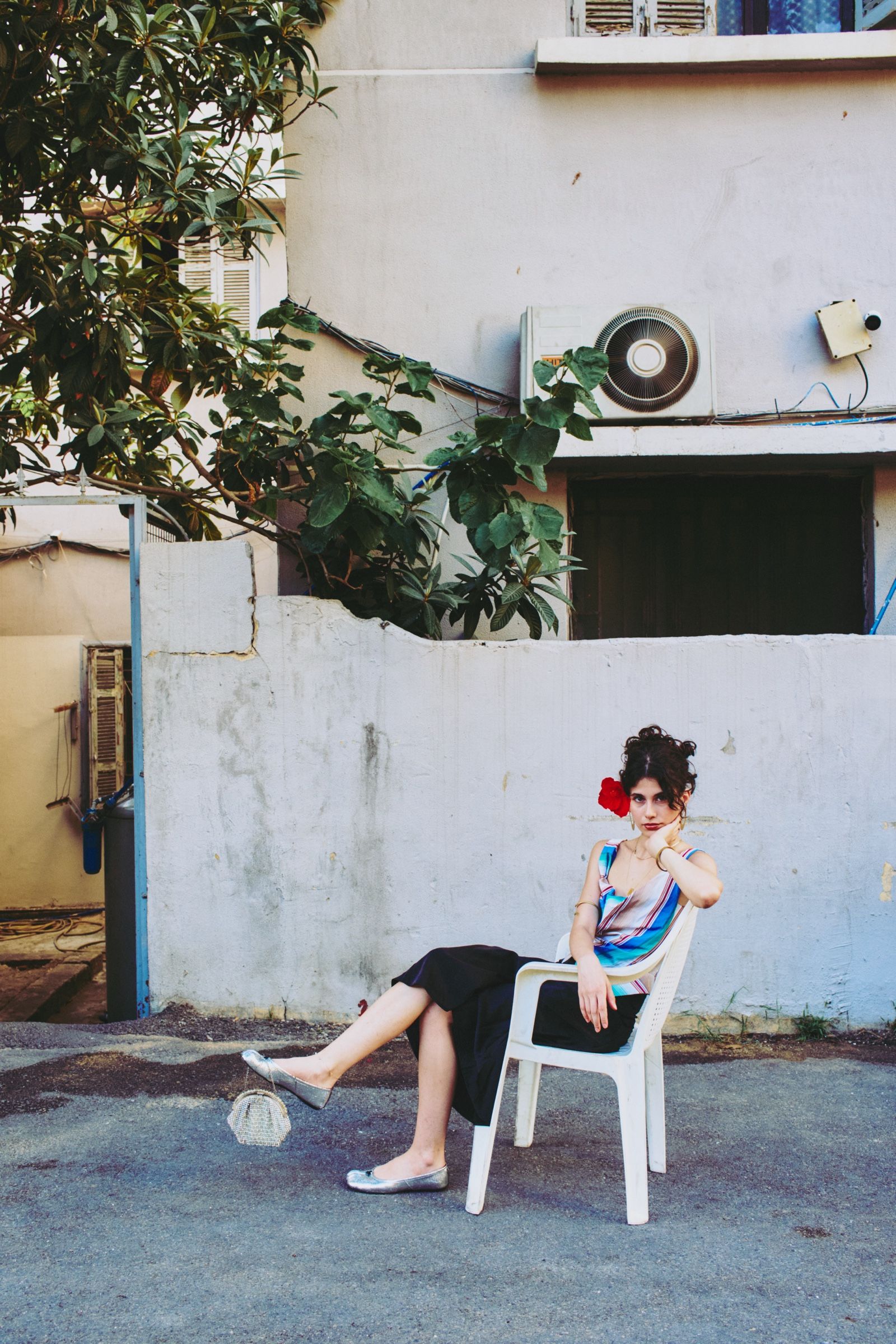 The model sits back in the chair on the tarmac, one leg stretched out, chin propped on her hand, looking straight at the camera.