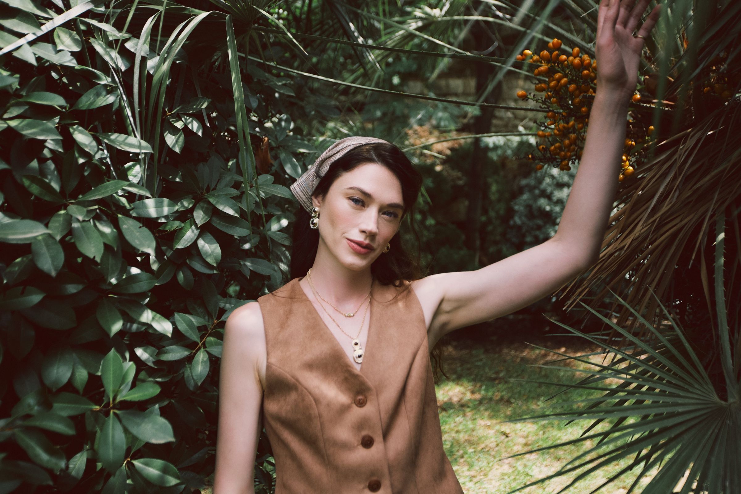 A model in a tan suede waistcoat and checked headscarf raises one arm to a palm frond, layered gold necklaces at the neck.