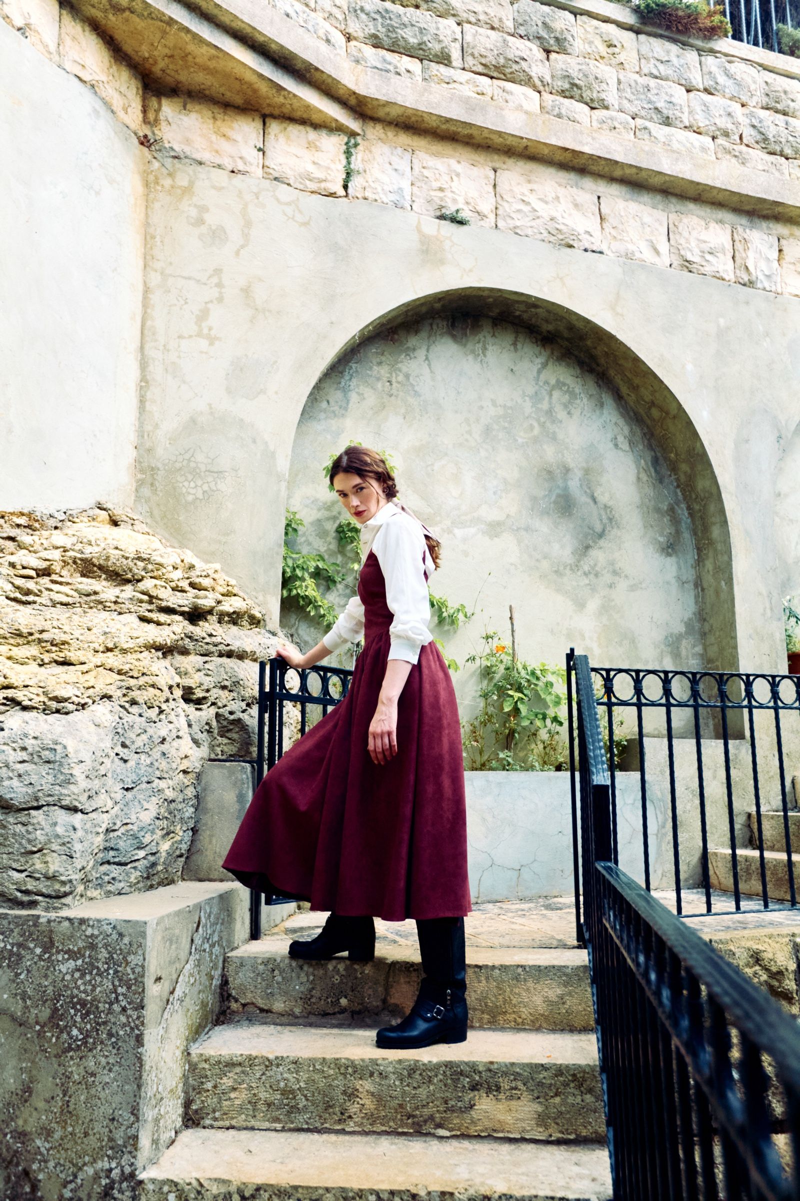 The model pauses on stone steps in a burgundy pinafore over a white blouse, looking back over her shoulder, one hand on an iron gate.