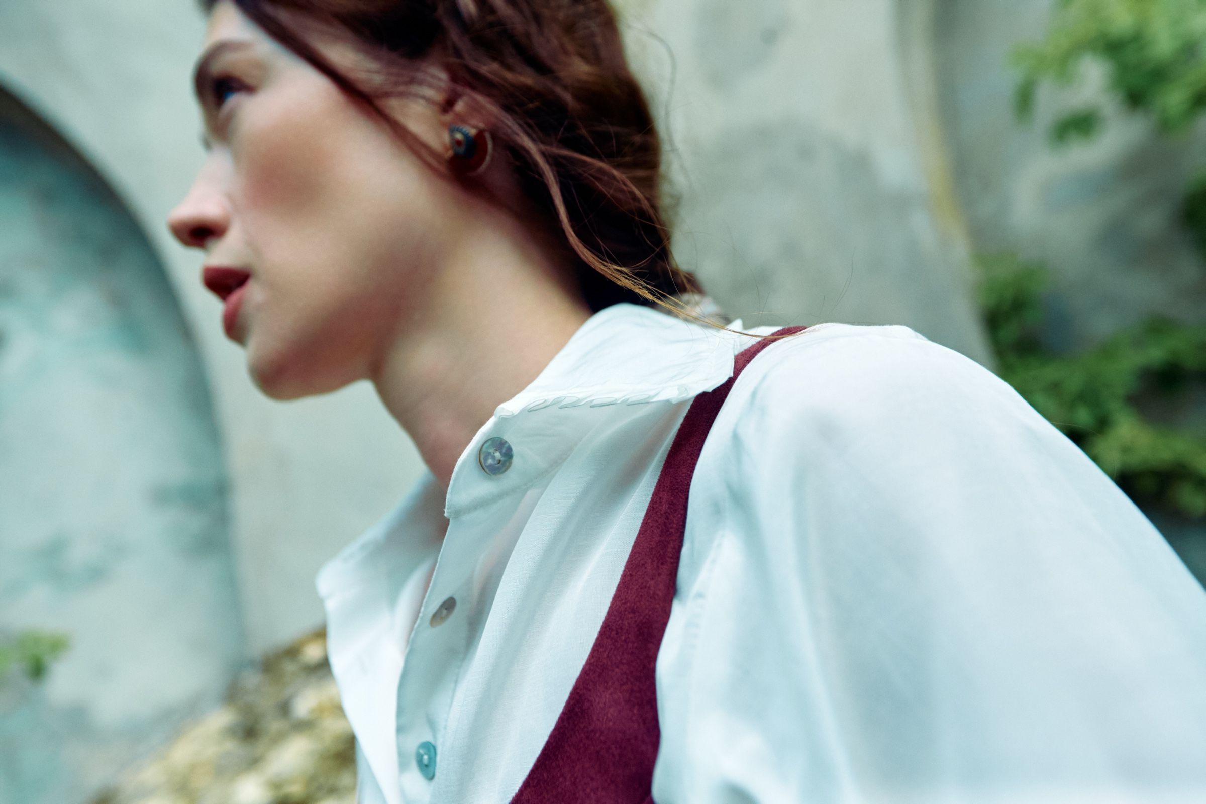 Close crop of a white collared blouse with shell buttons and a burgundy strap, the model turned in profile.
