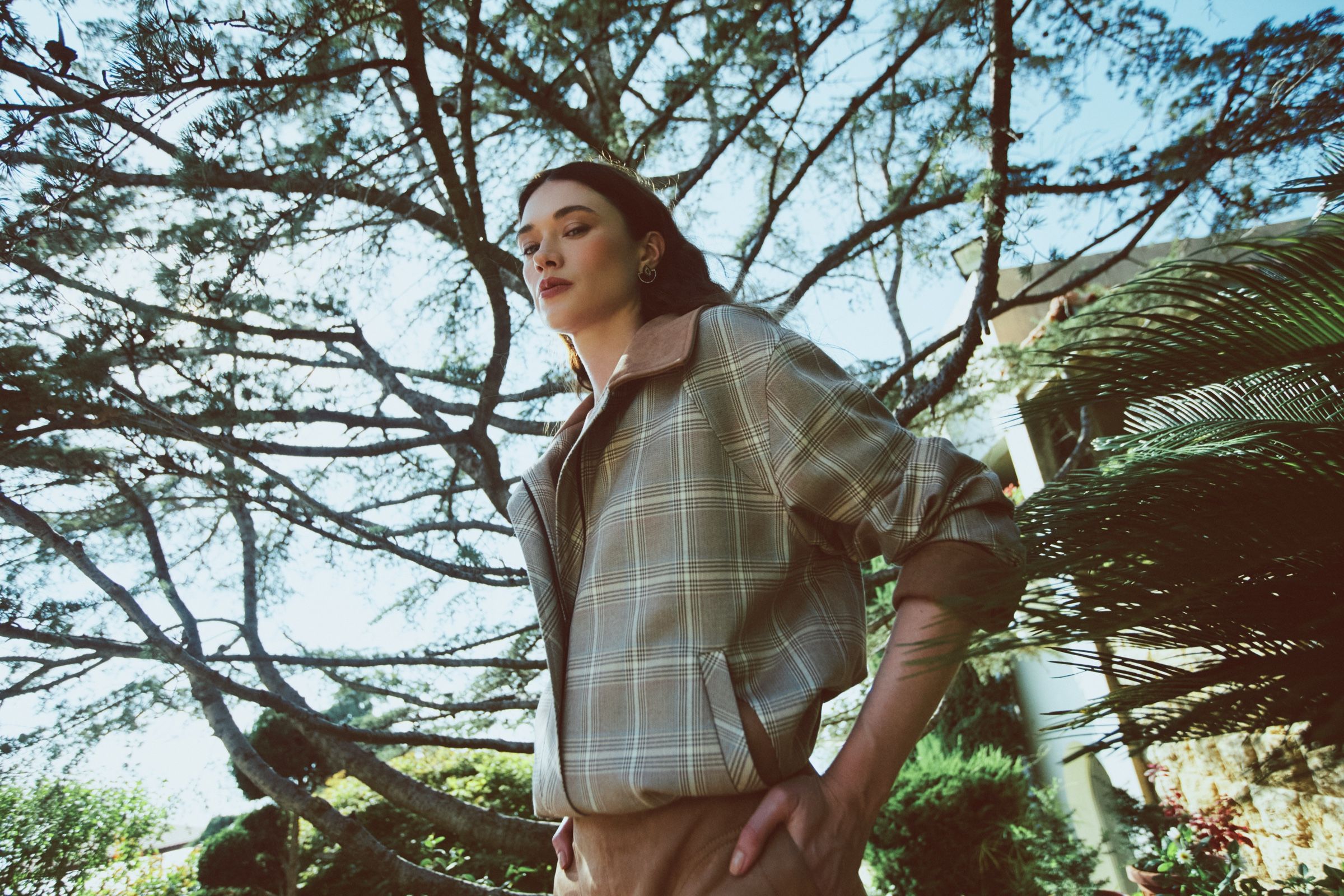 Low-angle view of a model in an oversized checked jacket with a suede collar, hand on hip against pine branches and bright sky.