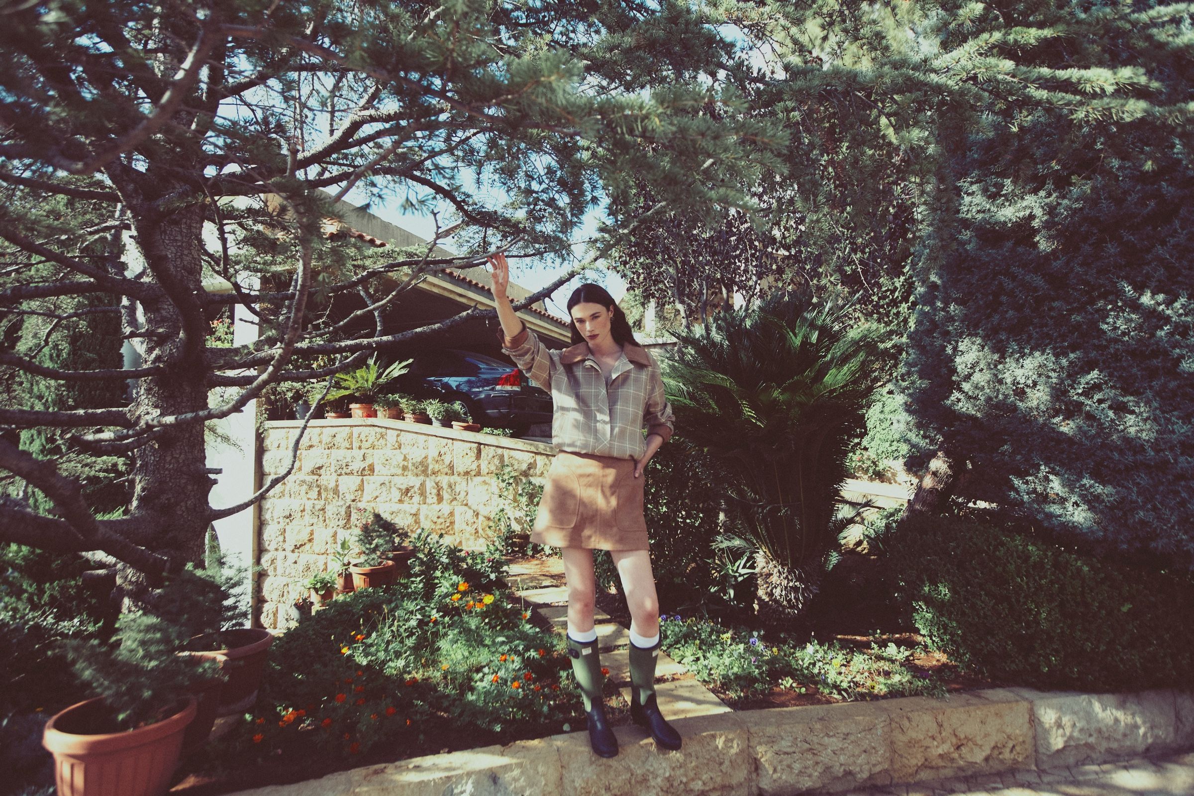 On a low garden wall, the model raises a hand to a pine branch in a checked jacket, tan suede miniskirt and olive rubber boots.