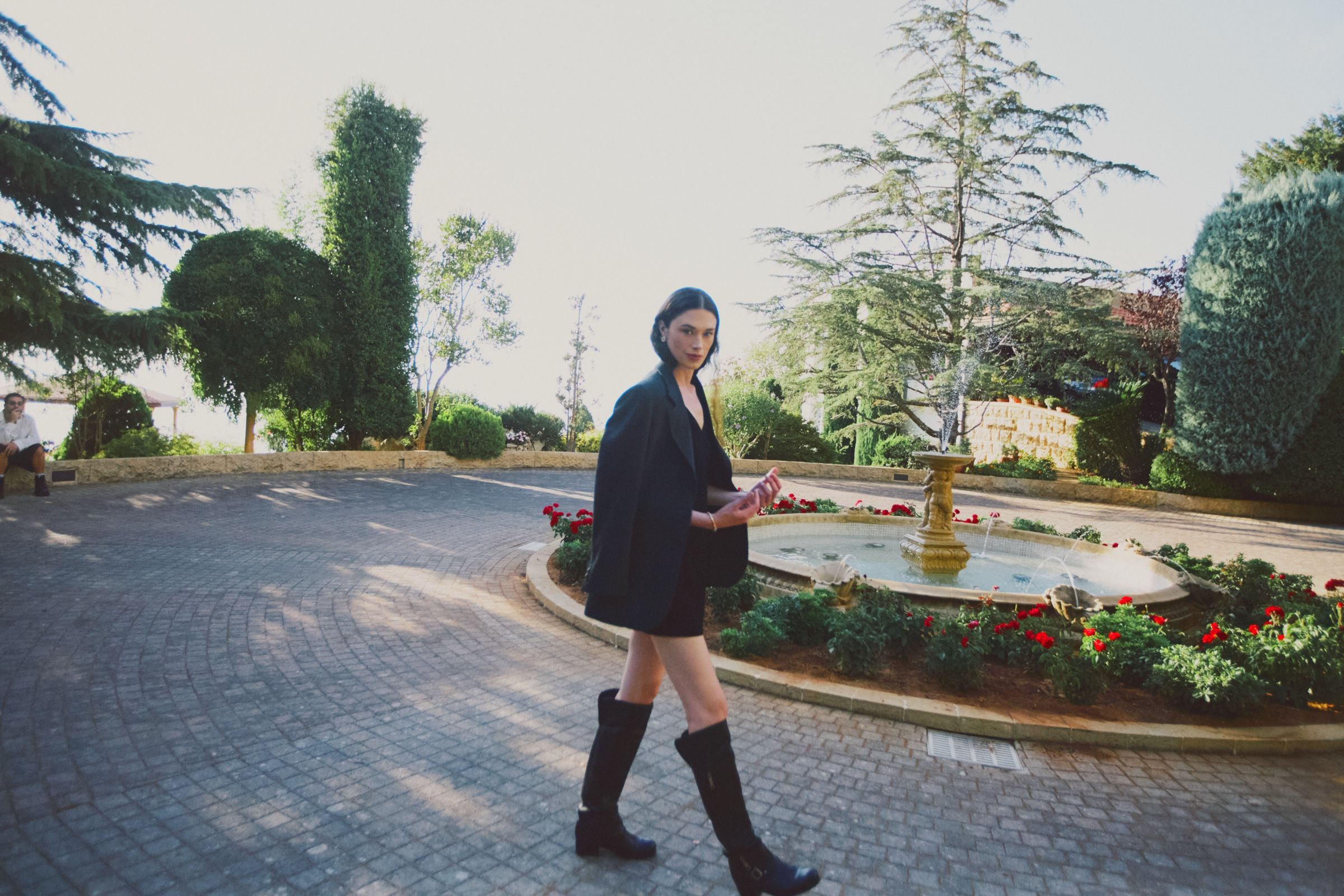 The model walks past a stone fountain and beds of red flowers in an oversized black blazer, short black dress and knee-high boots.