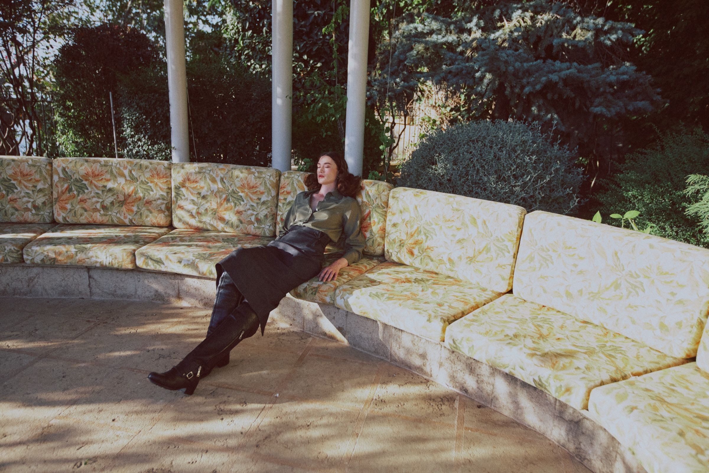 Head tipped back, eyes closed, the model rests along a leaf-print banquette in an olive satin shirt, dark leather skirt and tall black boots.