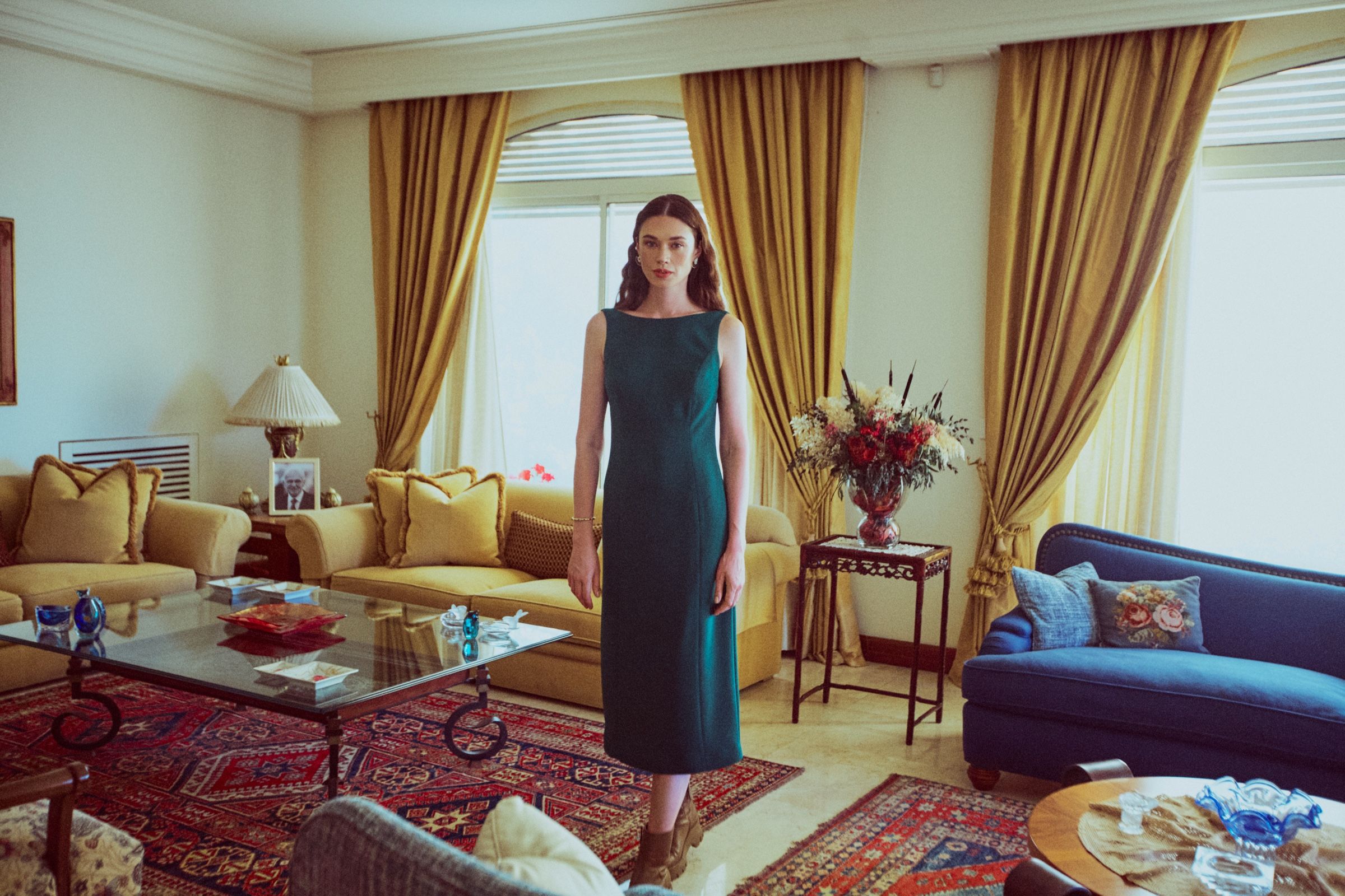 The model stands in a sunlit sitting room in a sleeveless dark green midi dress, gold curtains and patterned rugs around her.