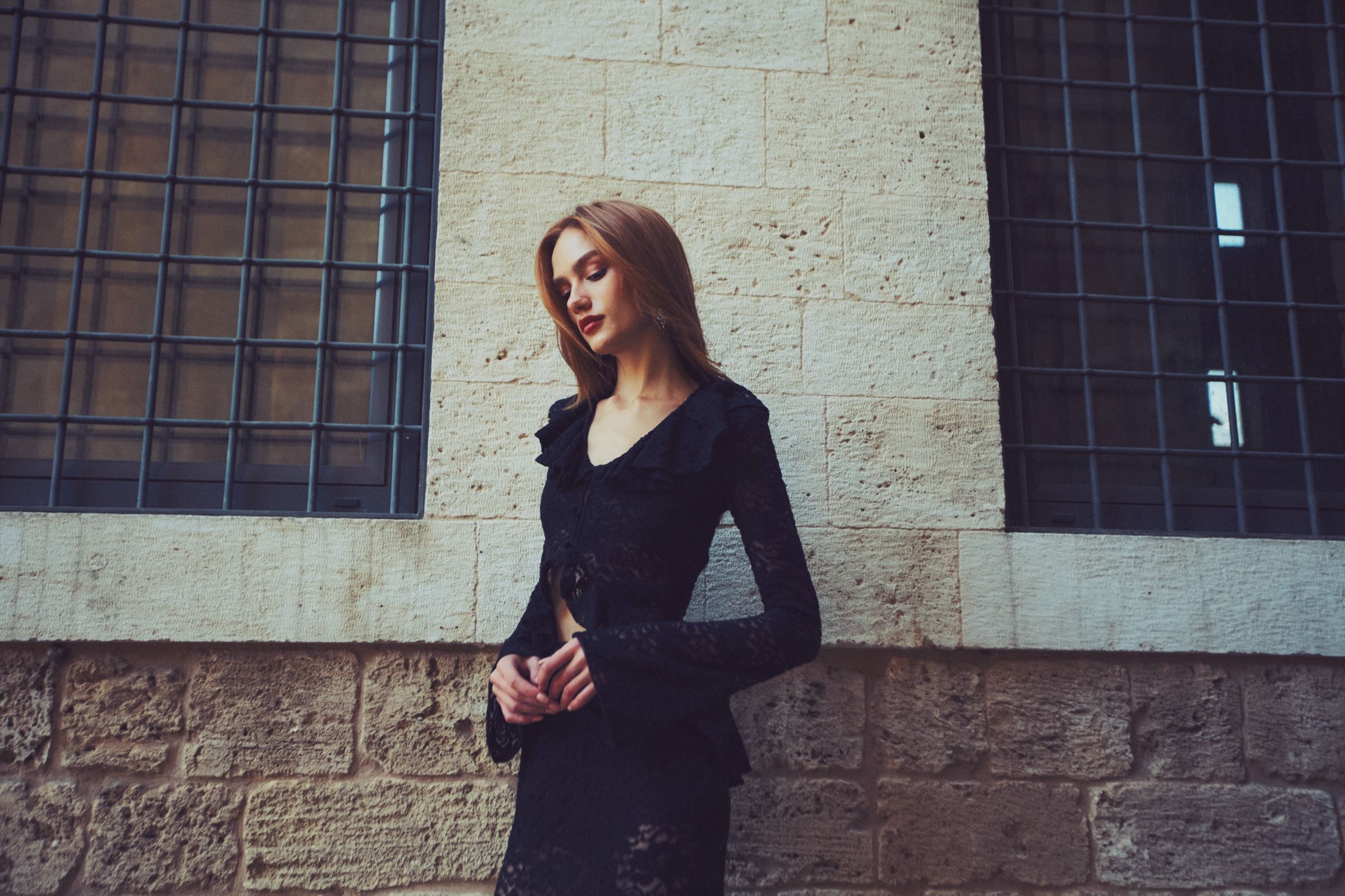 A model in a black lace top with flared sleeves stands with hands clasped before a limestone wall and barred windows.