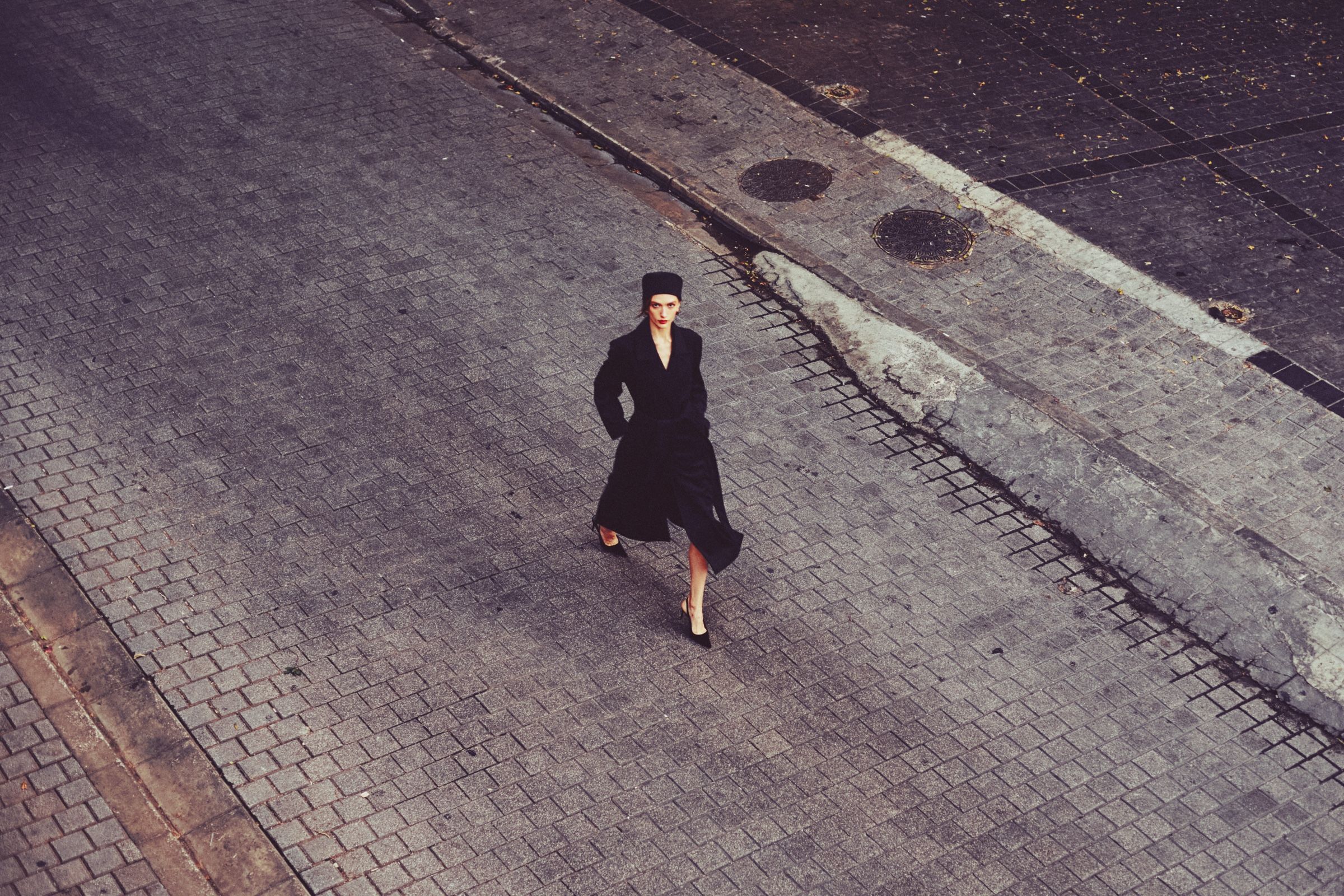 Shot from above, a woman in a long black coat and black hat walks across an empty paved street, looking up at the camera.