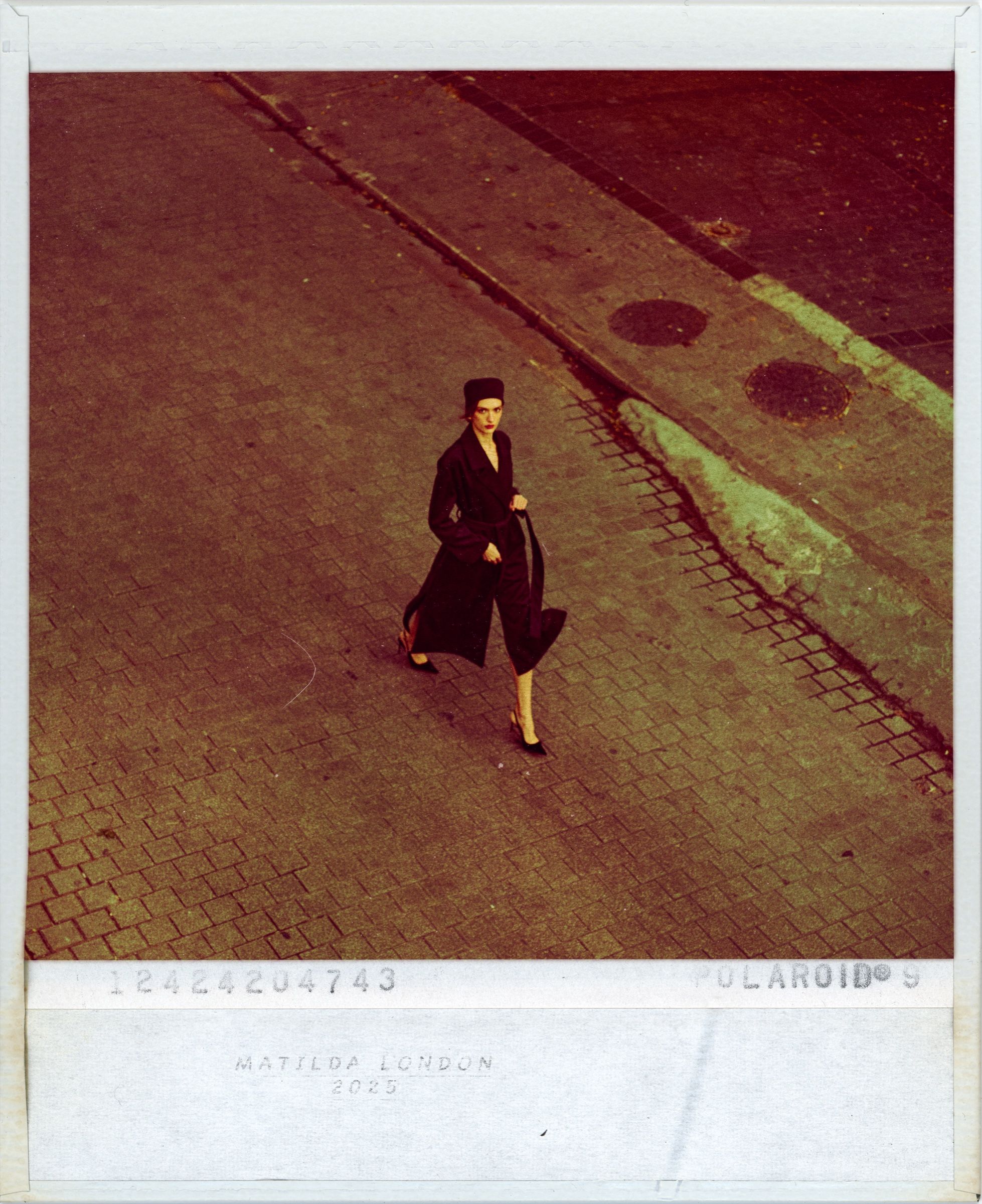 A Polaroid print, white border and all, of a woman in a black coat and hat crossing empty pavement, red-green film cast.